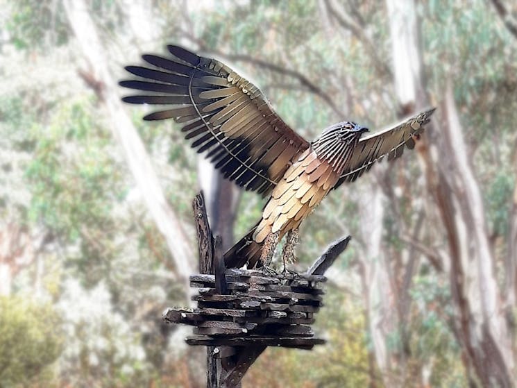 realistic metal sculpture of eagle in brown yellow tones with spread wings against forest background