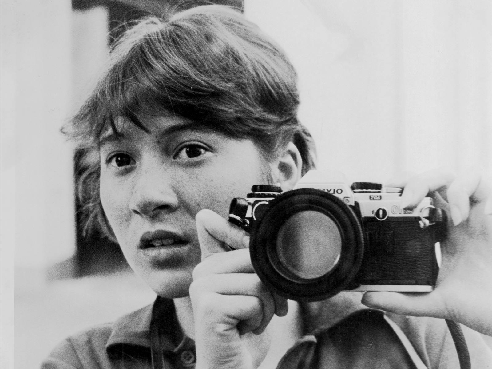 Jane Hutcheon holds a camera up to a mirror. She is young and has kind eyes.  The image is b&w.