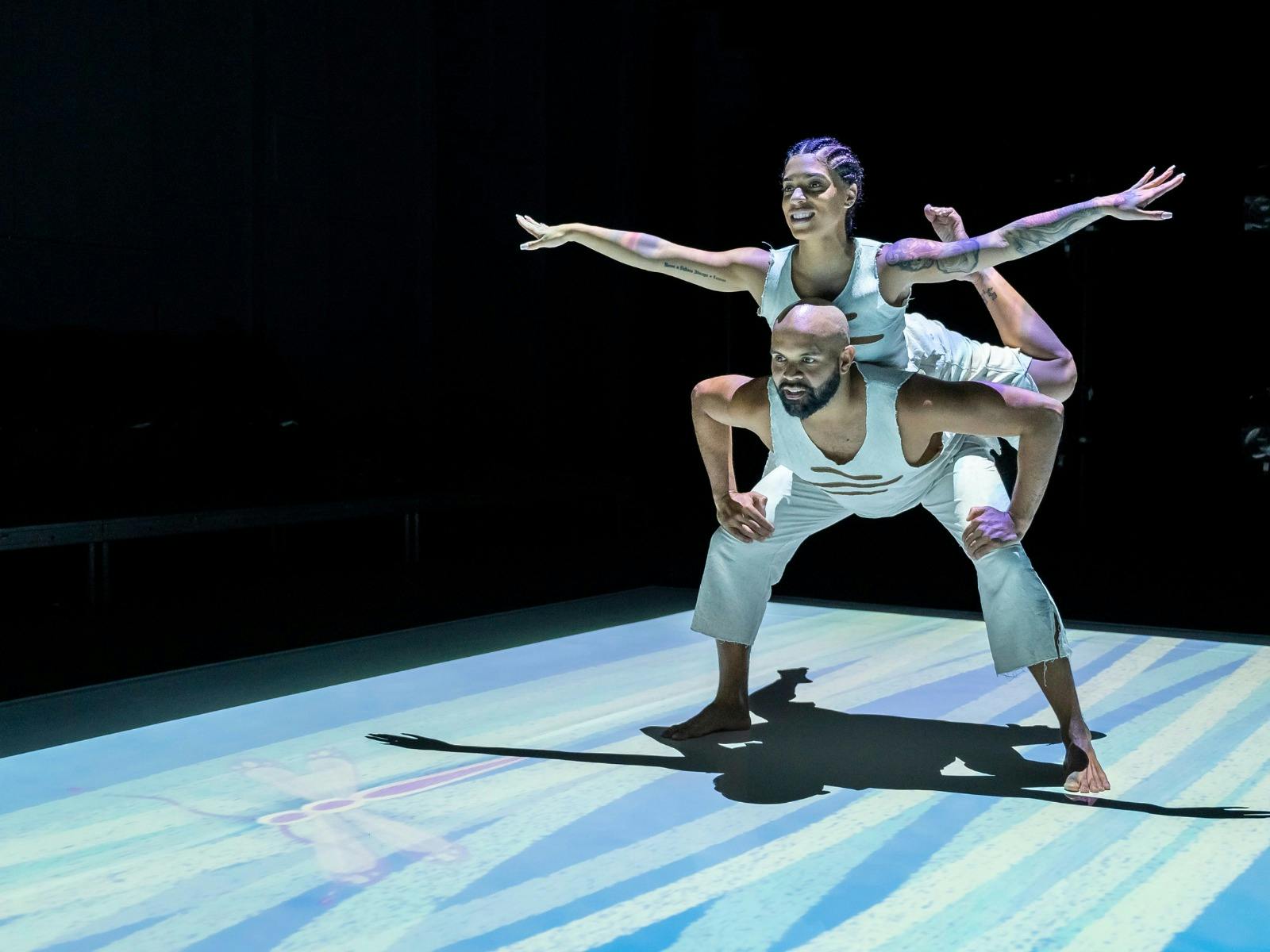 A woman balancing on the back of a man with her arms spread. They are lit by a projection.
