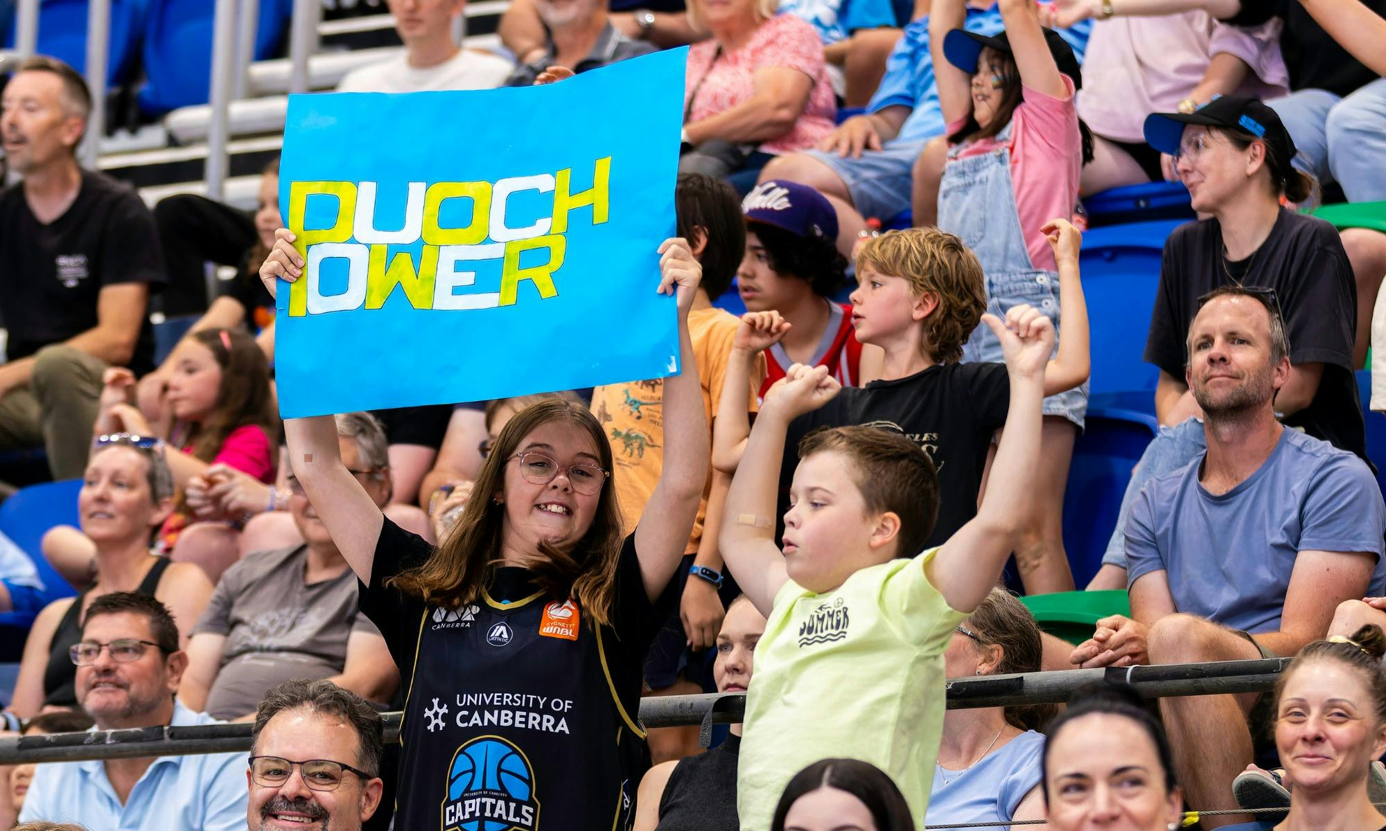 Two young fans cheering in the stands while waving a UC Capitals poster.