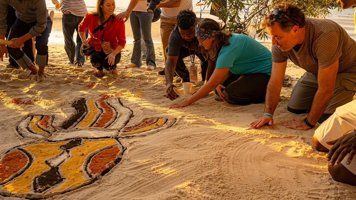 Butchulla man creating coloured sand art on a beach with a group of visitors on K'gari