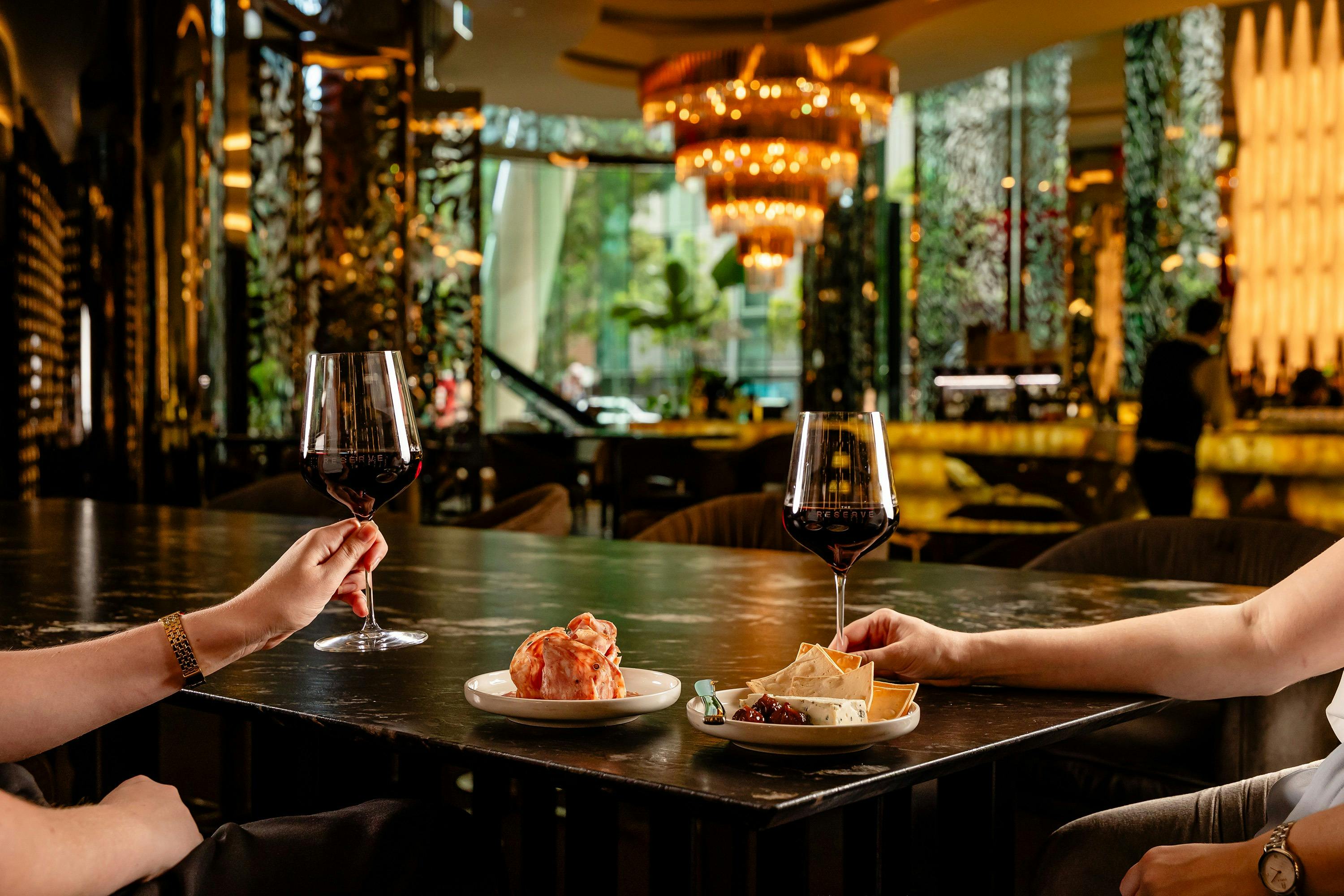 Warm interiors of The Reserve Wine Bar with two hands holding two red wine glasses and charcuterie.