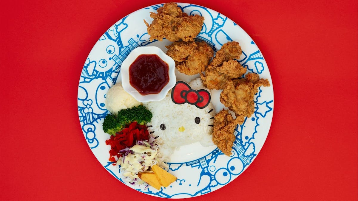 A Hello Kitty-shaped rice ball served with fried chicken, vegetables, and sauce on a plate.