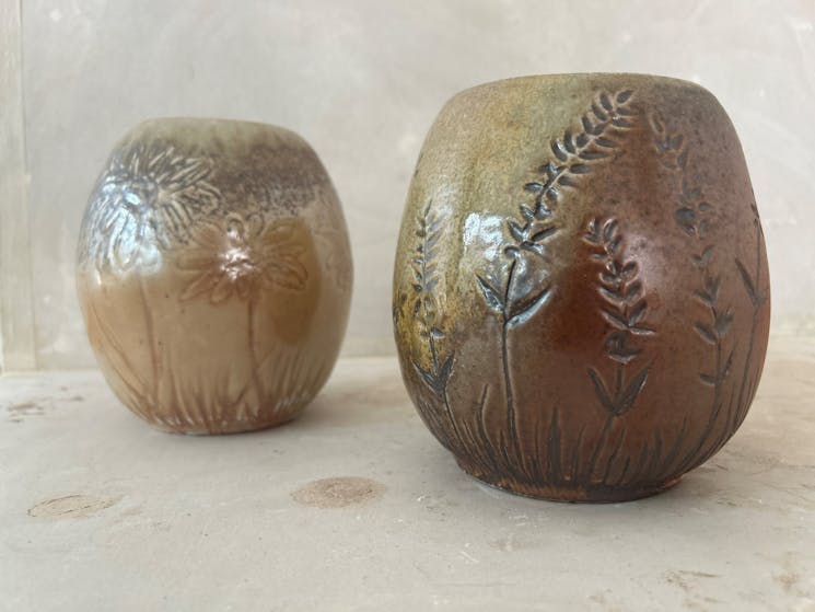 Two brown pots, cream surface. Left one has flowers carved, right one has flowering grass carving