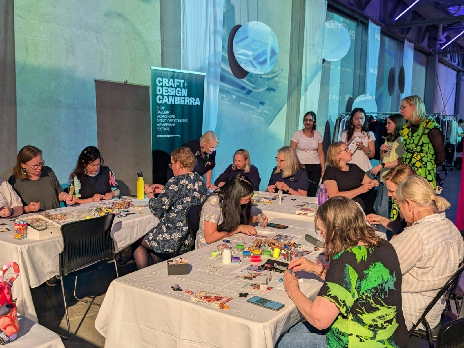 At a u-shaped table, a group of people sit and create small wove tokens out of recycled materials.