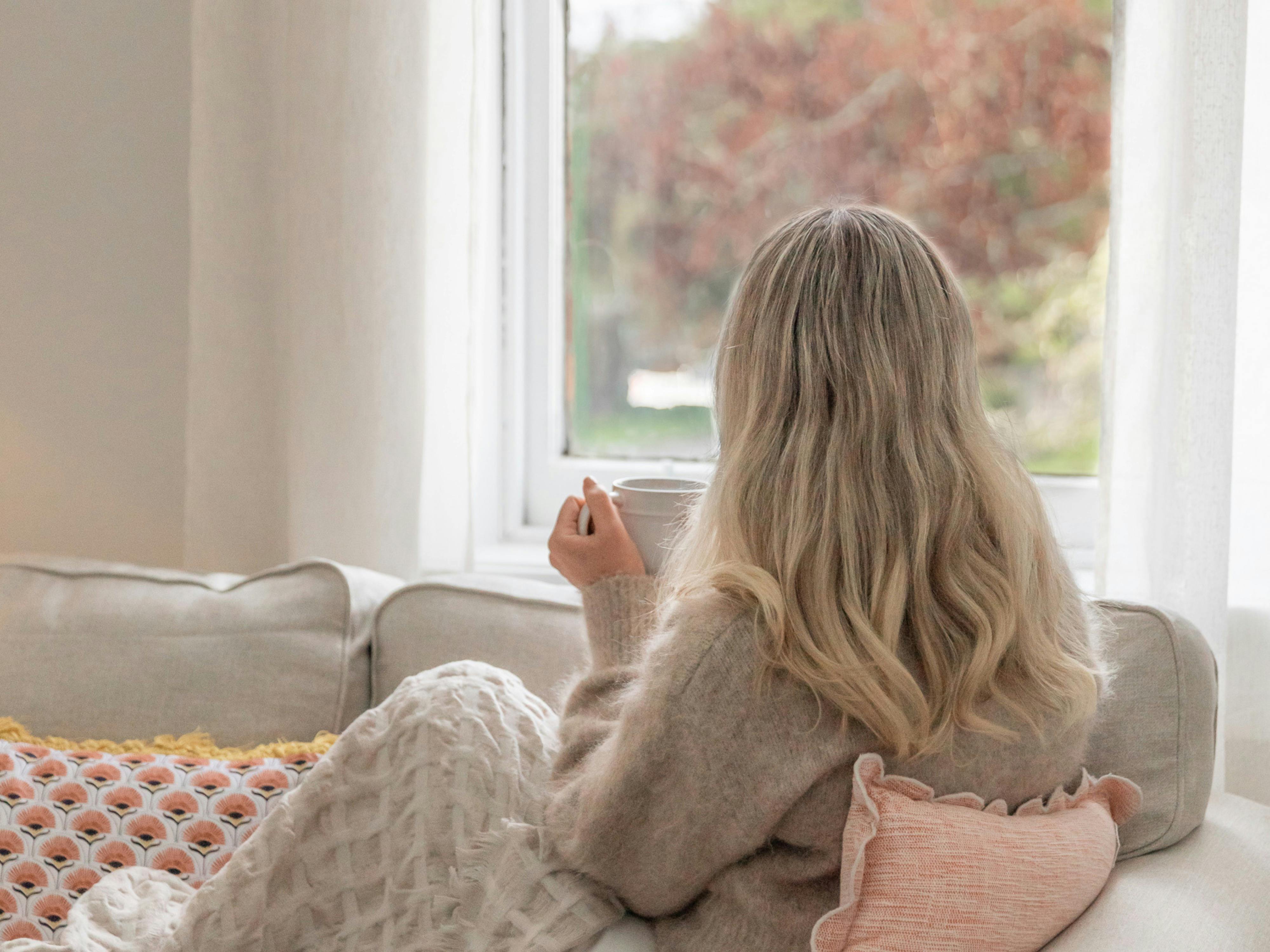 Woman sitting on cream sofa, holding hot drink, rug over legs, looking out window over city park
