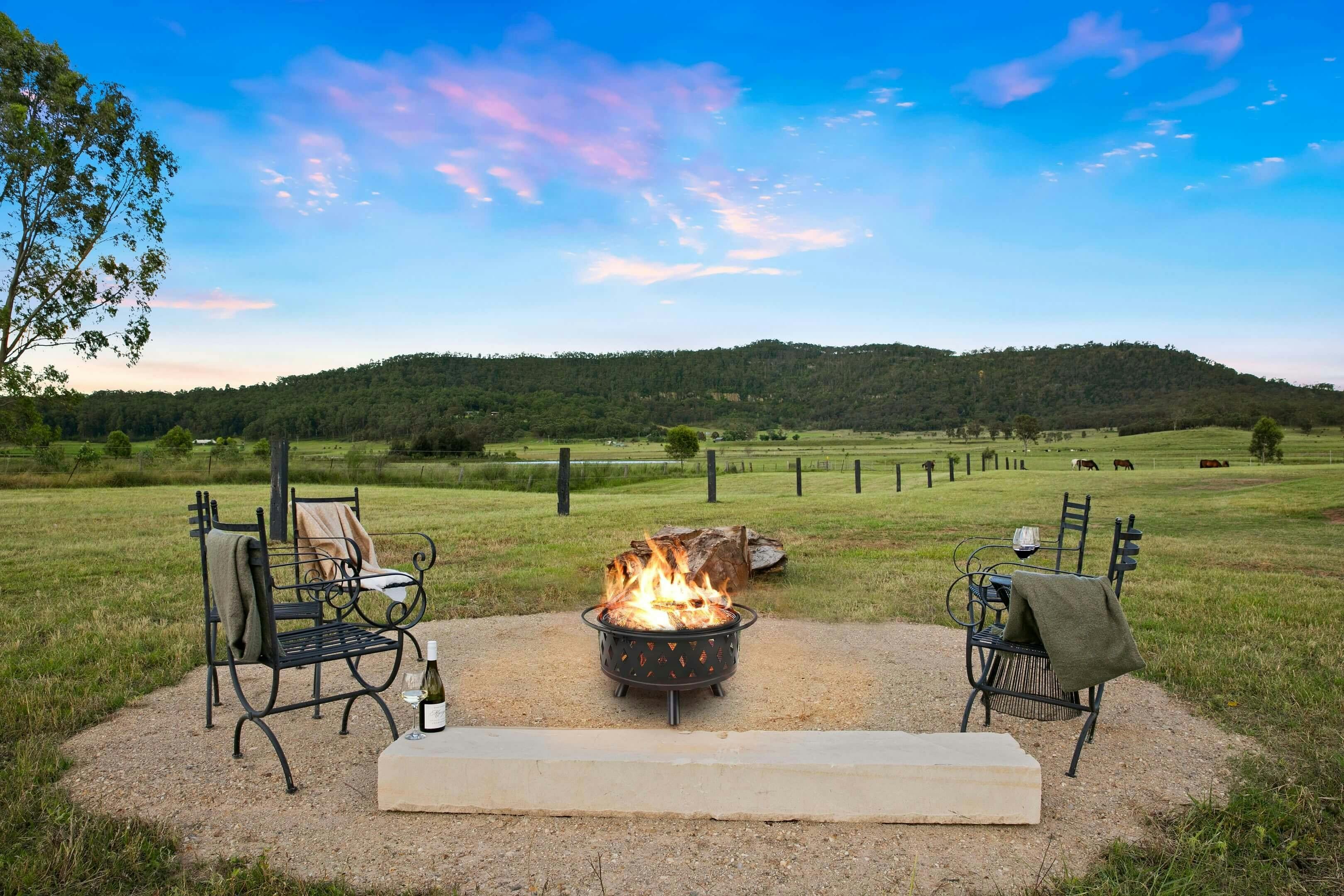 firepit on a patio in a grassy field at sunset, with wine bottlestwo chairs, a log bench, views