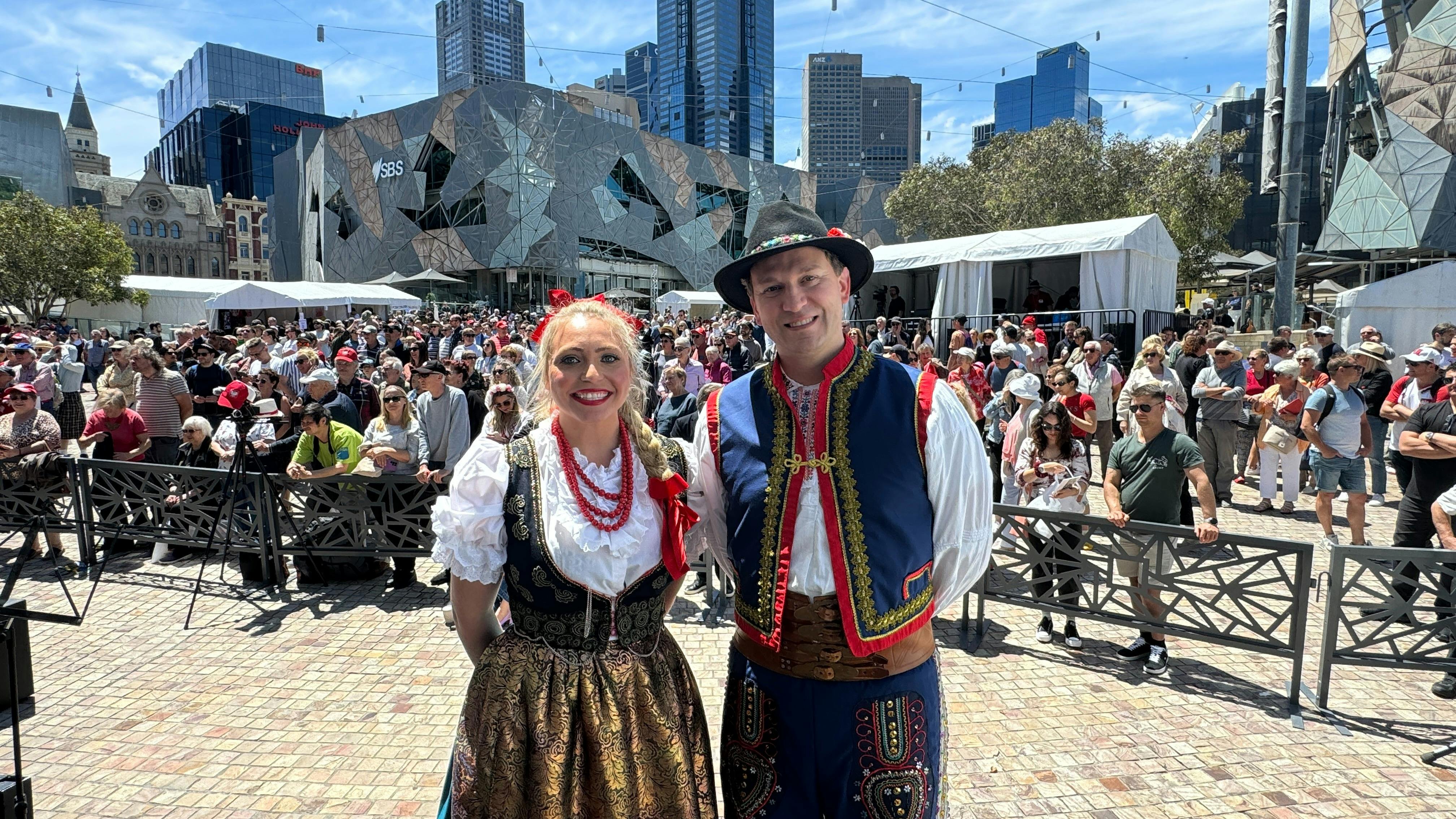 Two performers dressed in traditional Polish folk costumes smile for the camera in front of a large