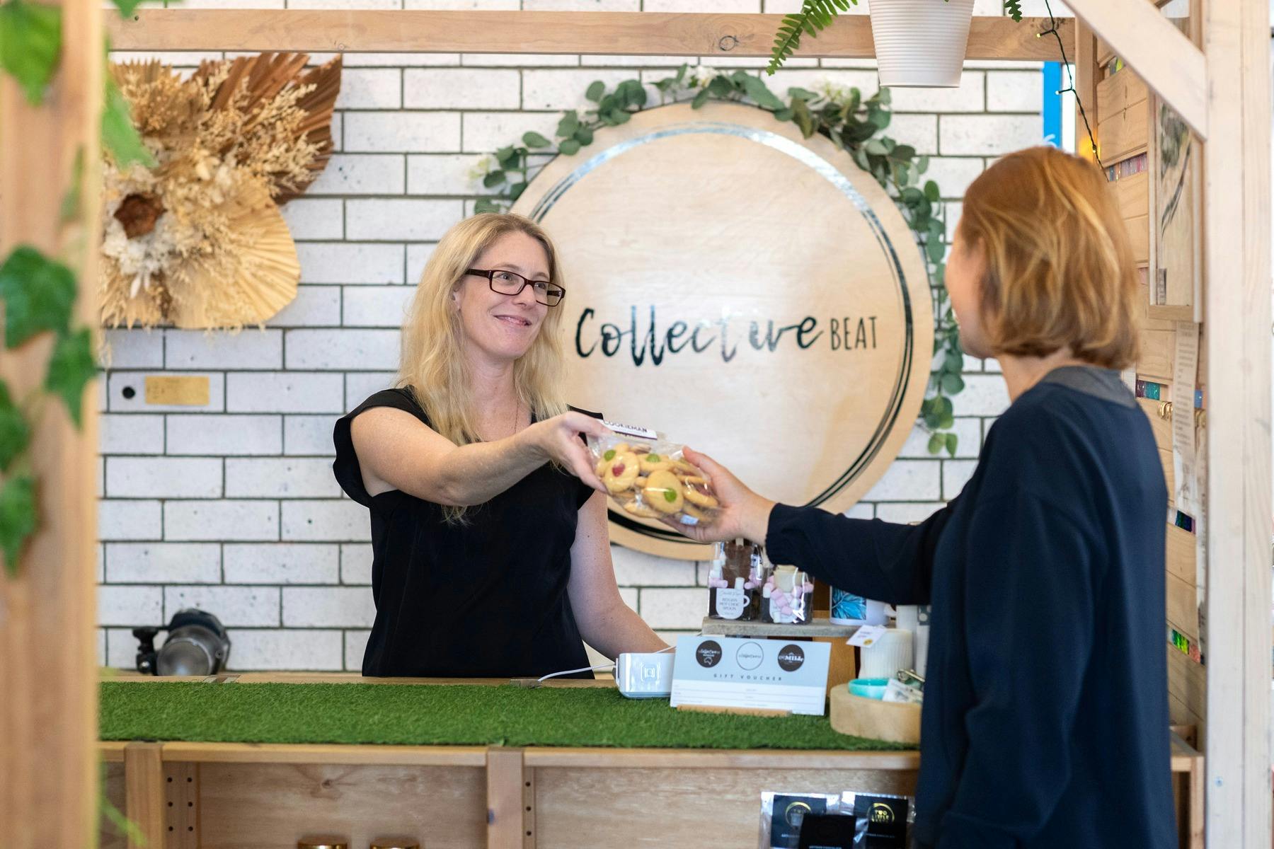 카운터 뒤에 있는 여성이 고객에게 제품을 건네주고 있습니다. 두 여성 모두 검은색 옷을 입고 있습니다.