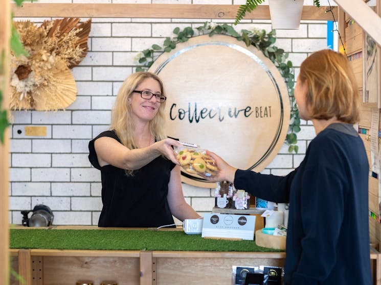 Woman behind counter, handing product to a customer. both women wearing black