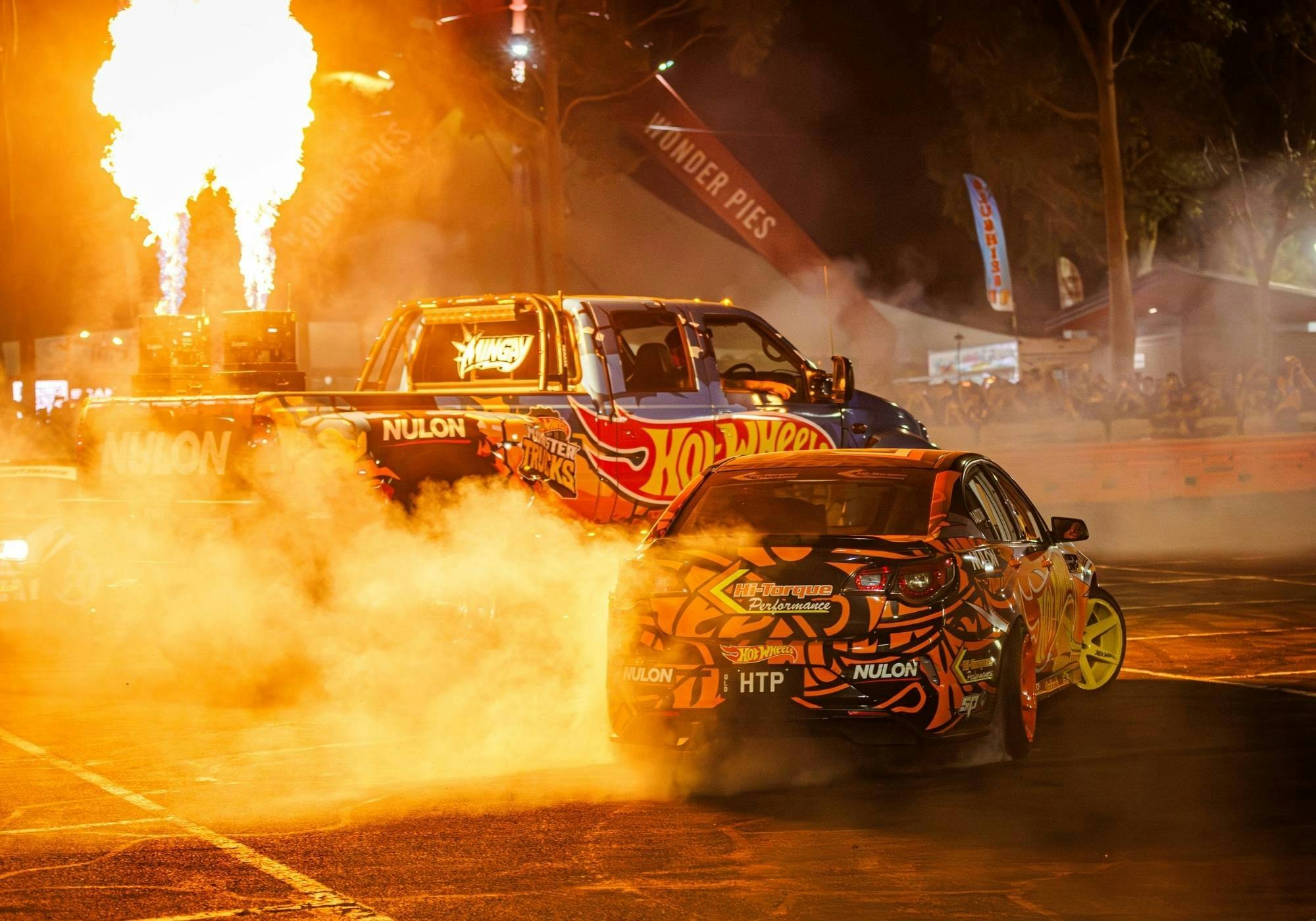 Hot Wheels-liveried performance vehicles producing tyre smoke beside large flame effects at night.