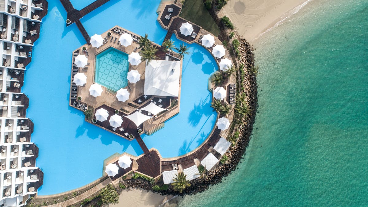 Aerial shot of a hexagonal pool near a sandy beach with clear turquoise water and palm trees.