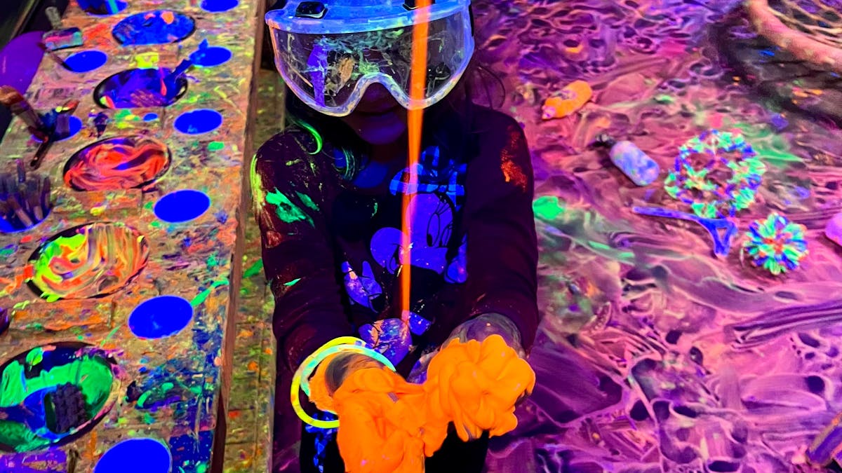 child with goggles on catching pouring glowing paint at Messy Glow Co UV Paint Room