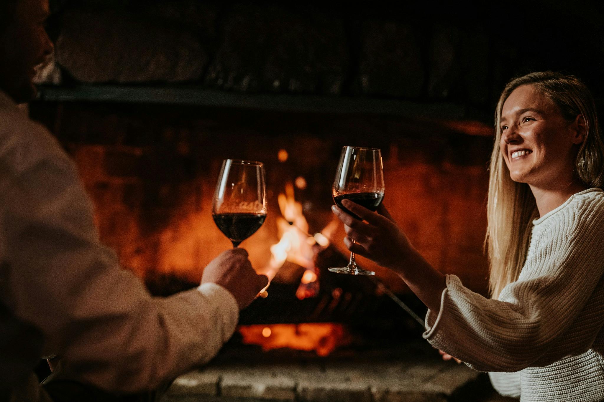 A couple cheers red wine by an open fire with content smiling expressions.