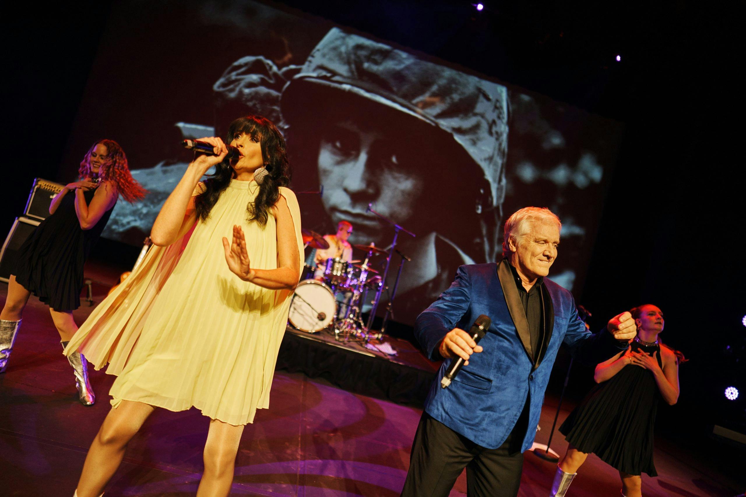 Two singers on stage in front of a projection of a vietnam war face.
