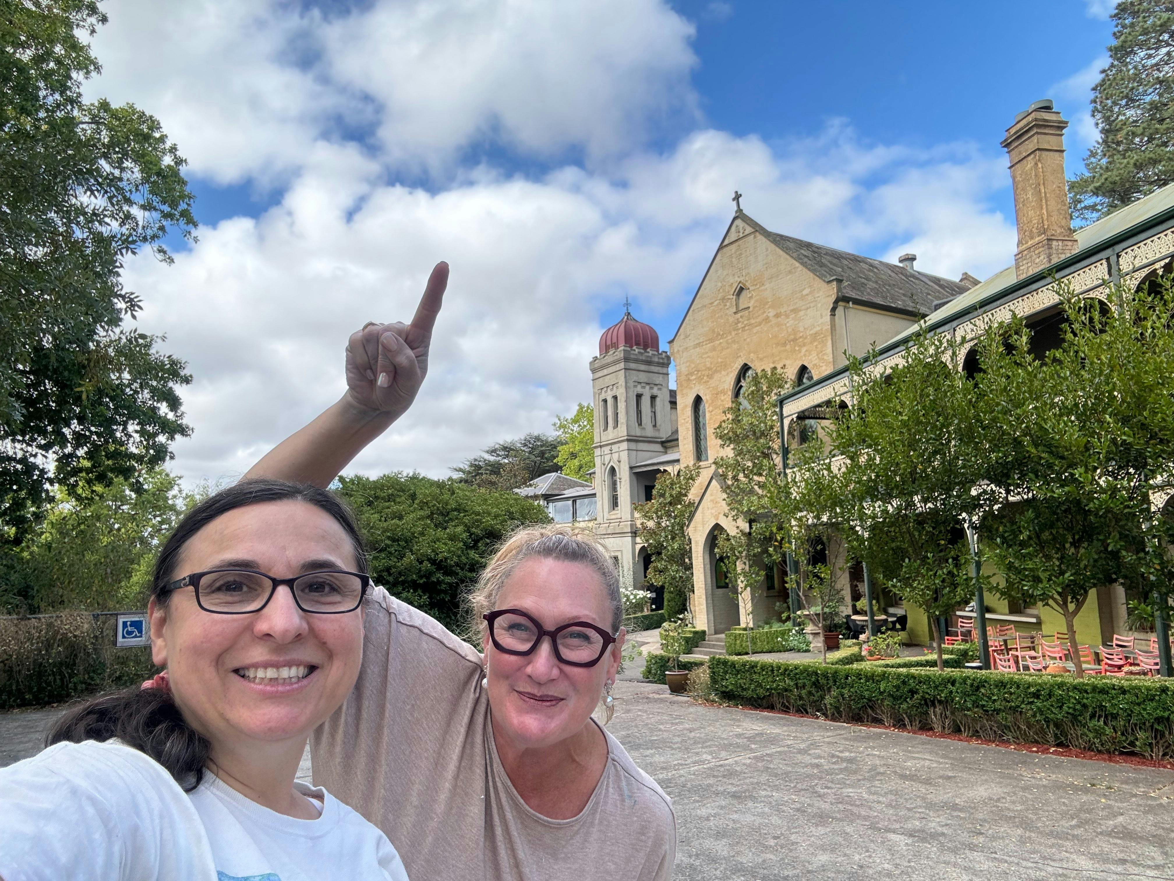 two women smiling at the camera with on pointing at the sky, with a brick convent in the background