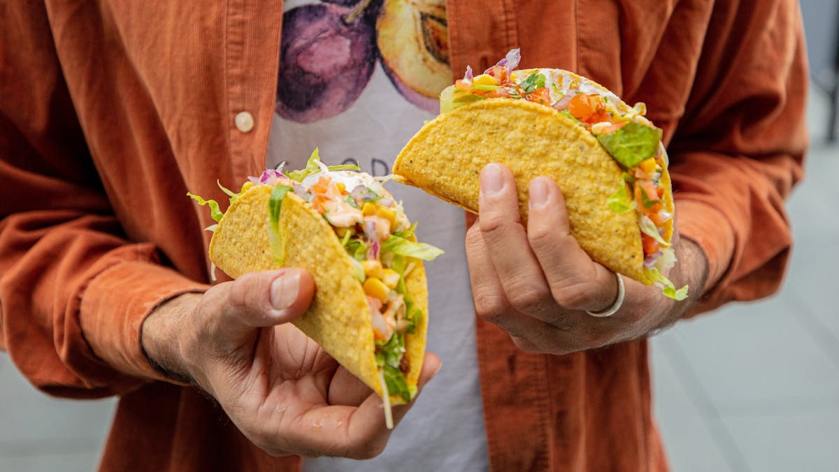 A person holding a hard shell taco in each hand, full with fillings and salsa, topped with sauce