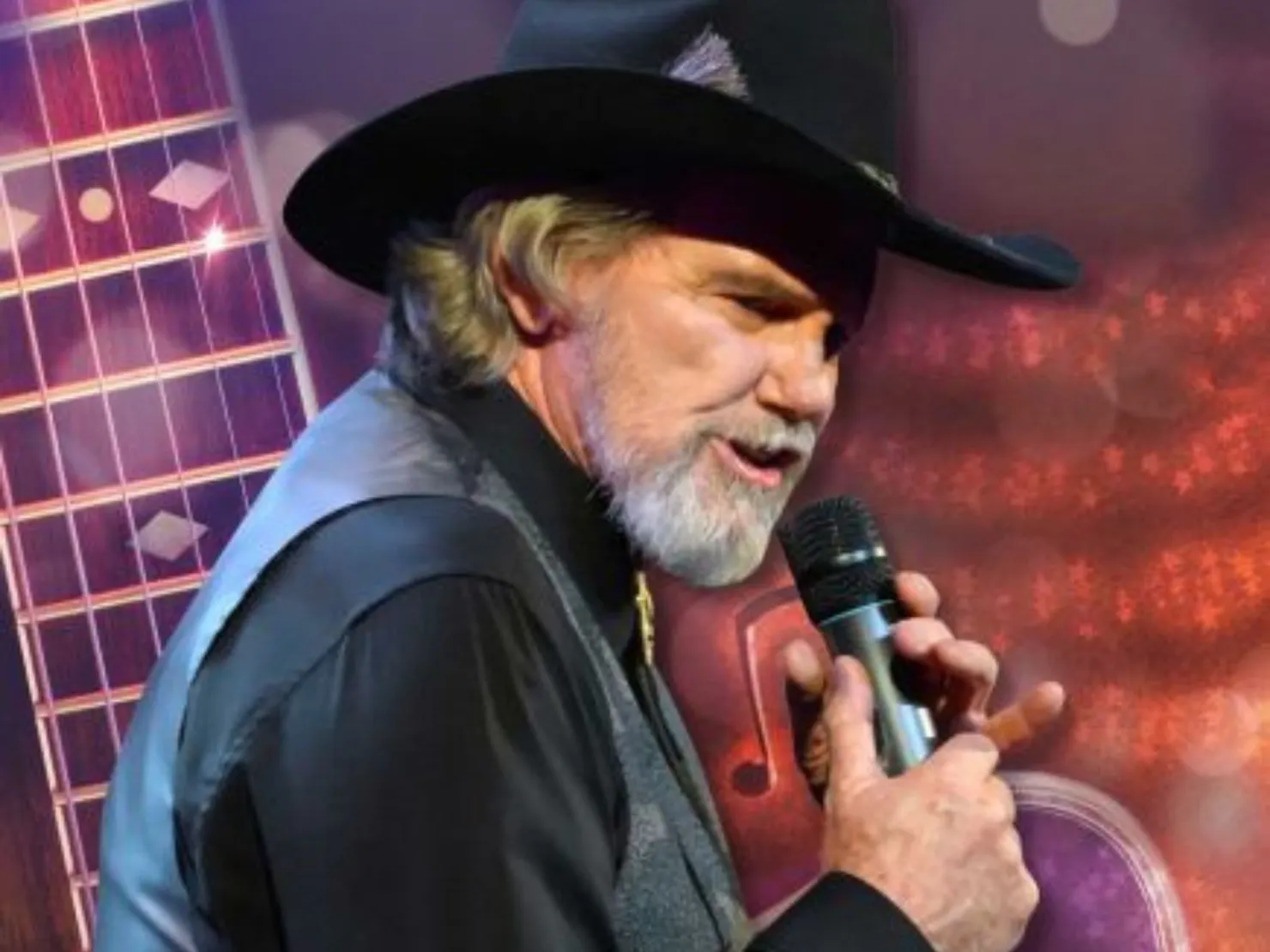 Man in black hat grey beard black shirt with vest with a microphone