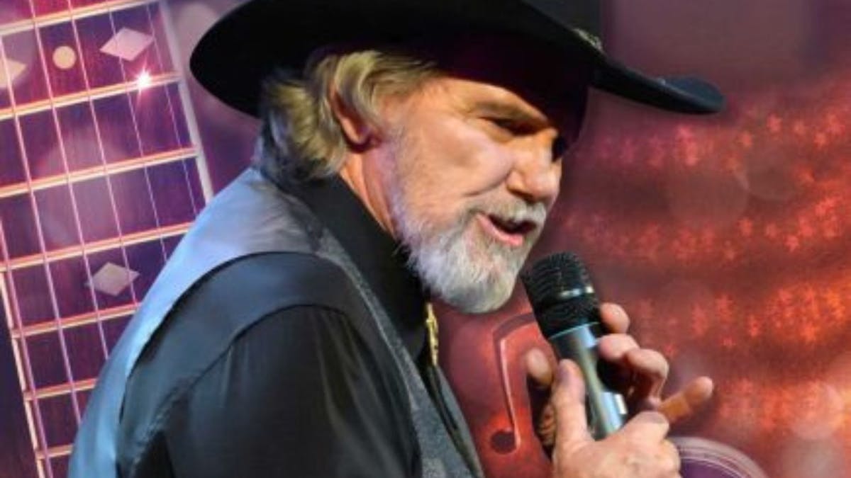 Man in black hat grey beard black shirt with vest with a microphone