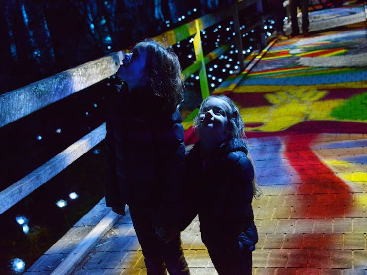 Two children standing on boardwalk with colourful projections in background