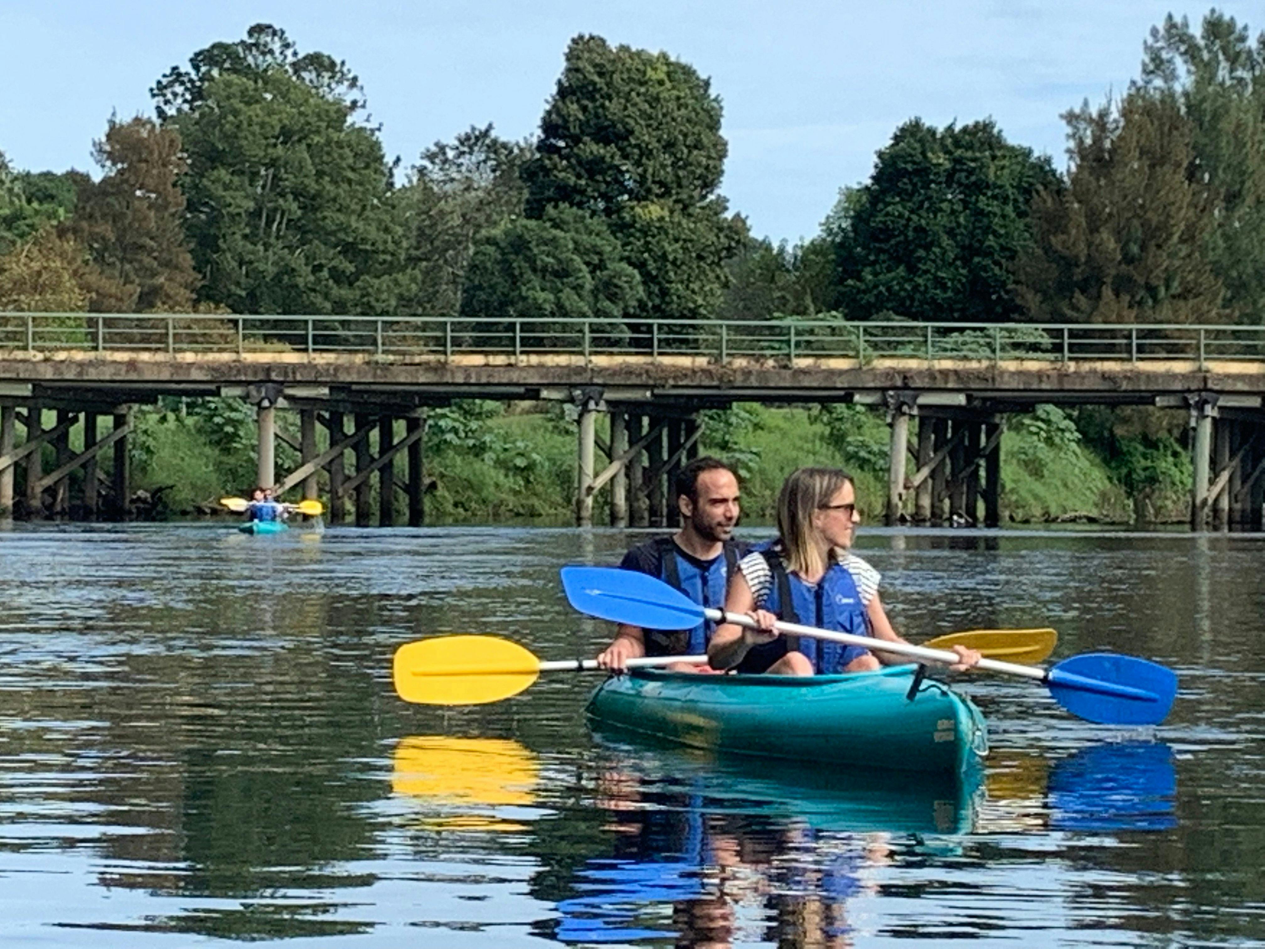 Man & woman sitting in a double canoe paddling in calm waters under a bridge with Bellingen Canoes