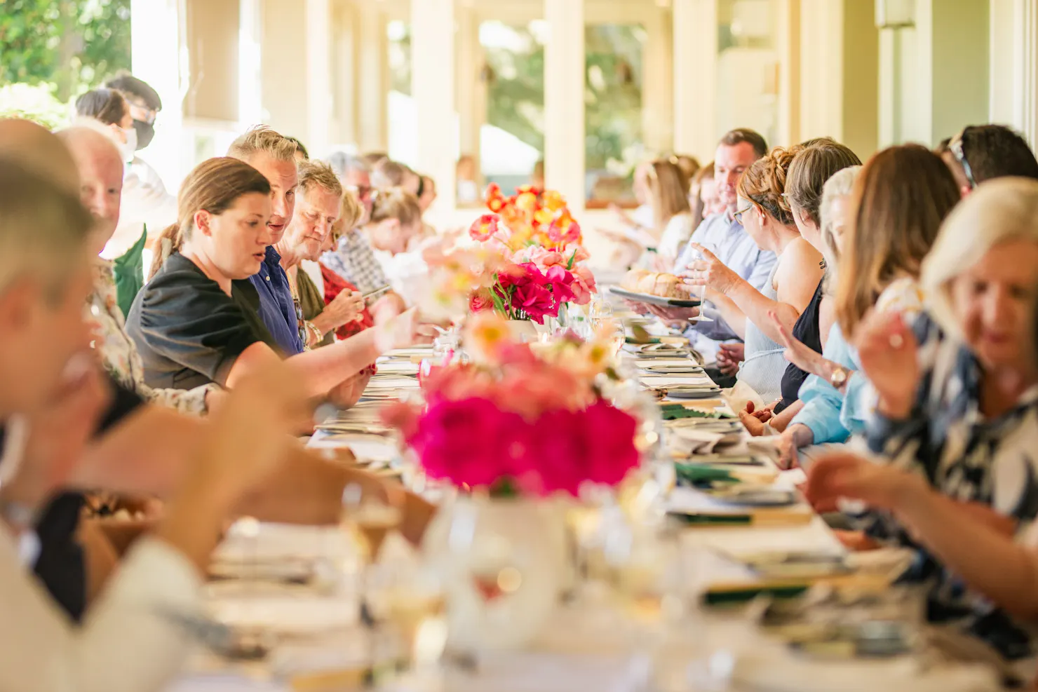 A long table full of people dining with flower decorations along the middle