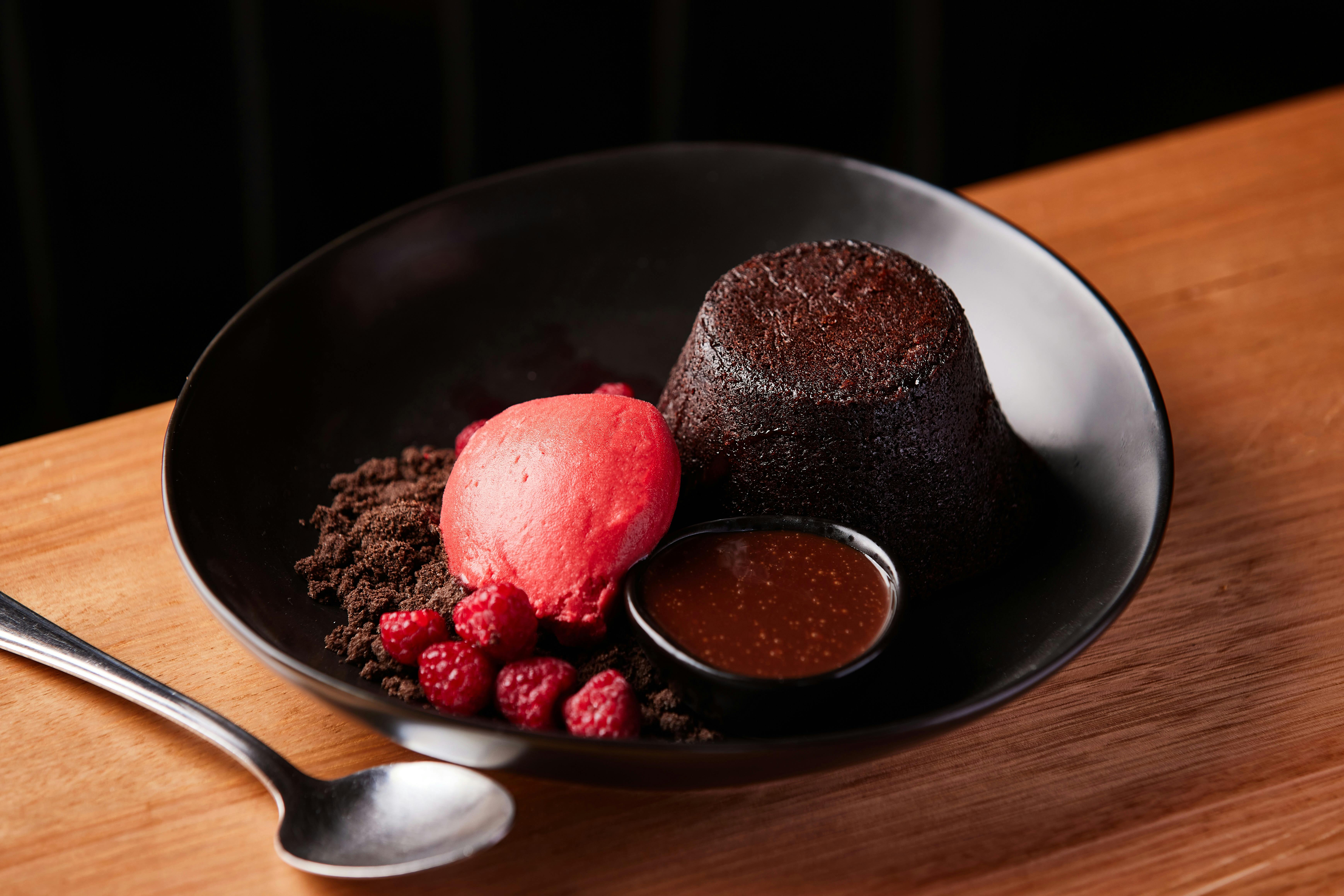 A chocolate lava pudding sits on a black plate with a pop of raspberry sorbet and dipping sauce