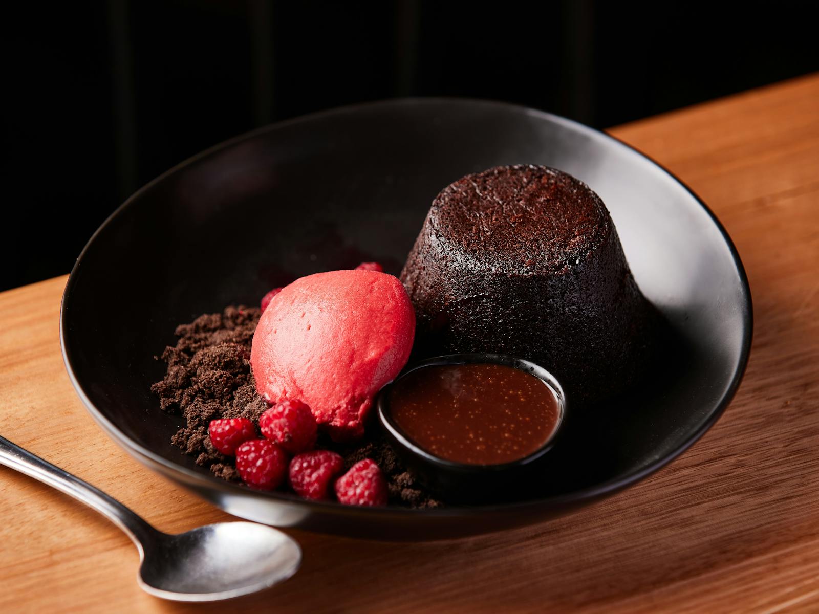 A chocolate lava pudding sits on a black plate with a pop of raspberry sorbet and dipping sauce