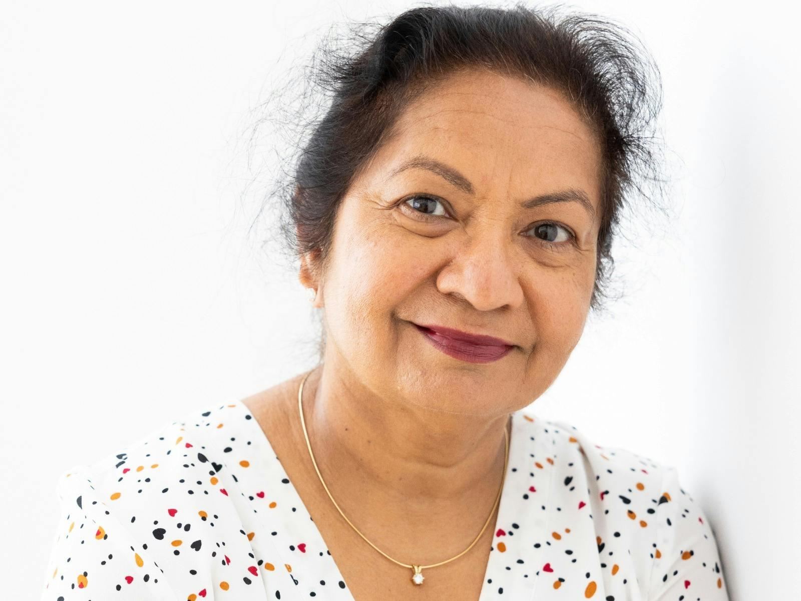 A woman smiling while resting her right hand on her left shoulder, while wearing a polka dot shirt.