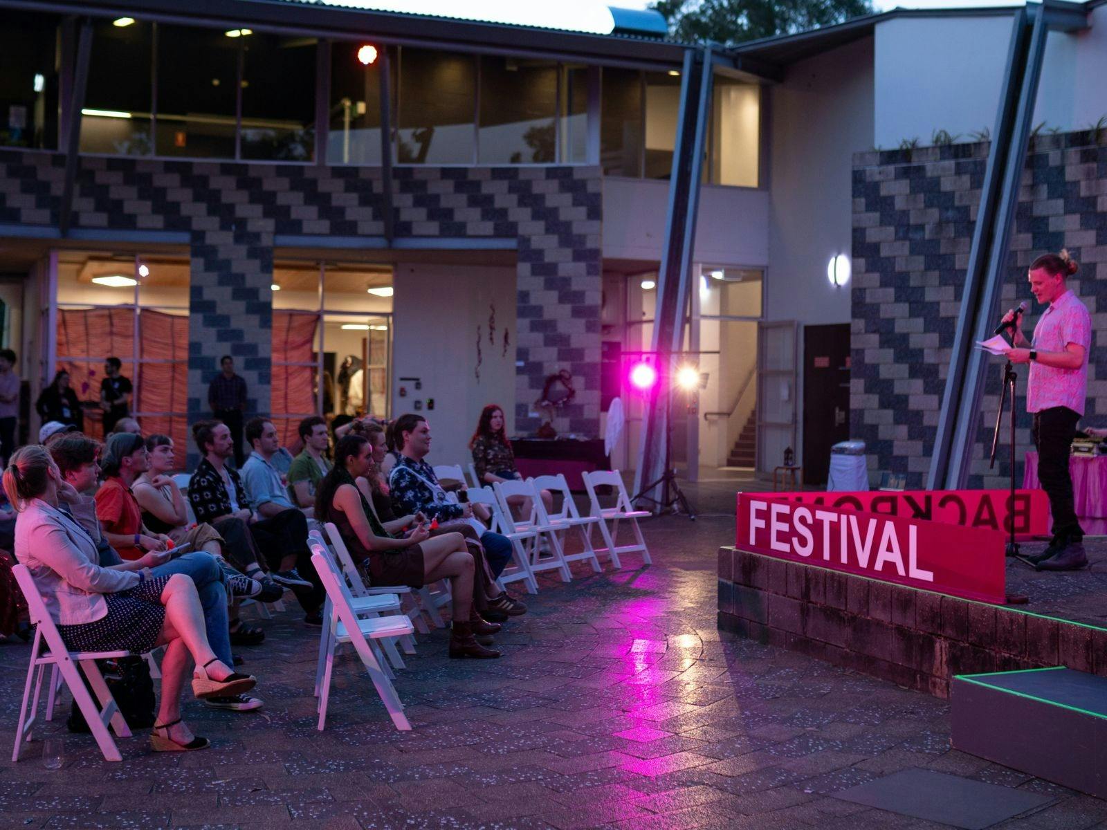 A crowd sitting on white chairs looking at a man giving a speech on stage