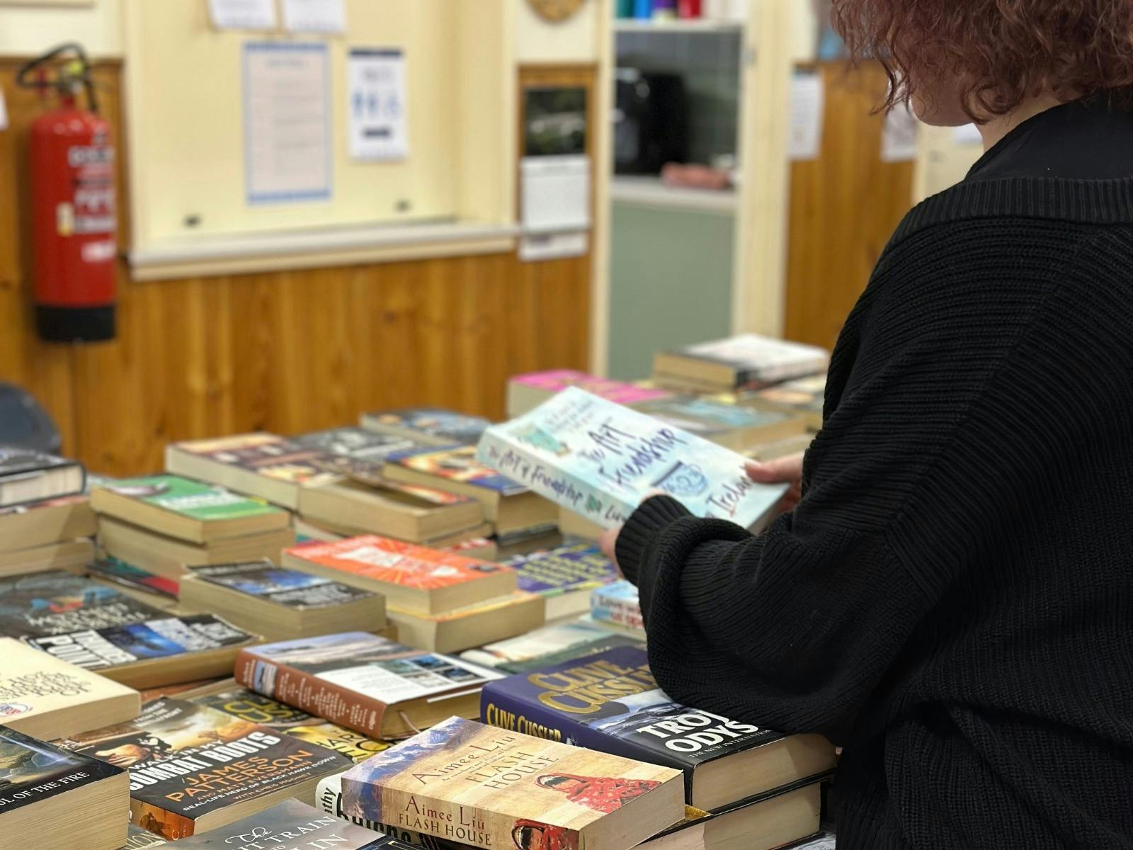 A person wearing a black cardigan holds a book while looking at a table stacked with books