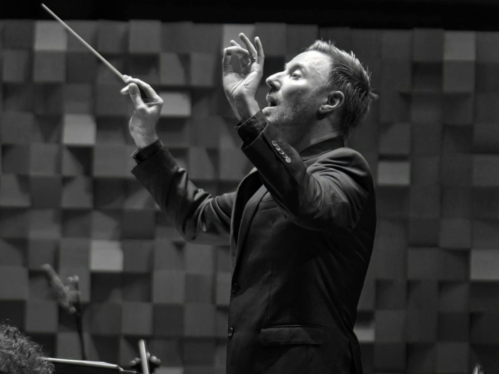 Monochrome image of a conductor, holding a baton, with his hands in the air.