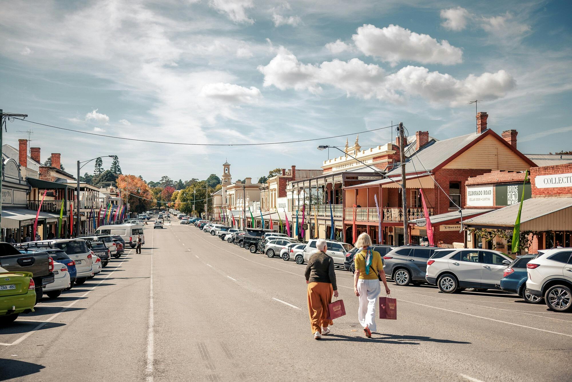 Two women with shopping bags walking down a historic street with colourful banners and cars