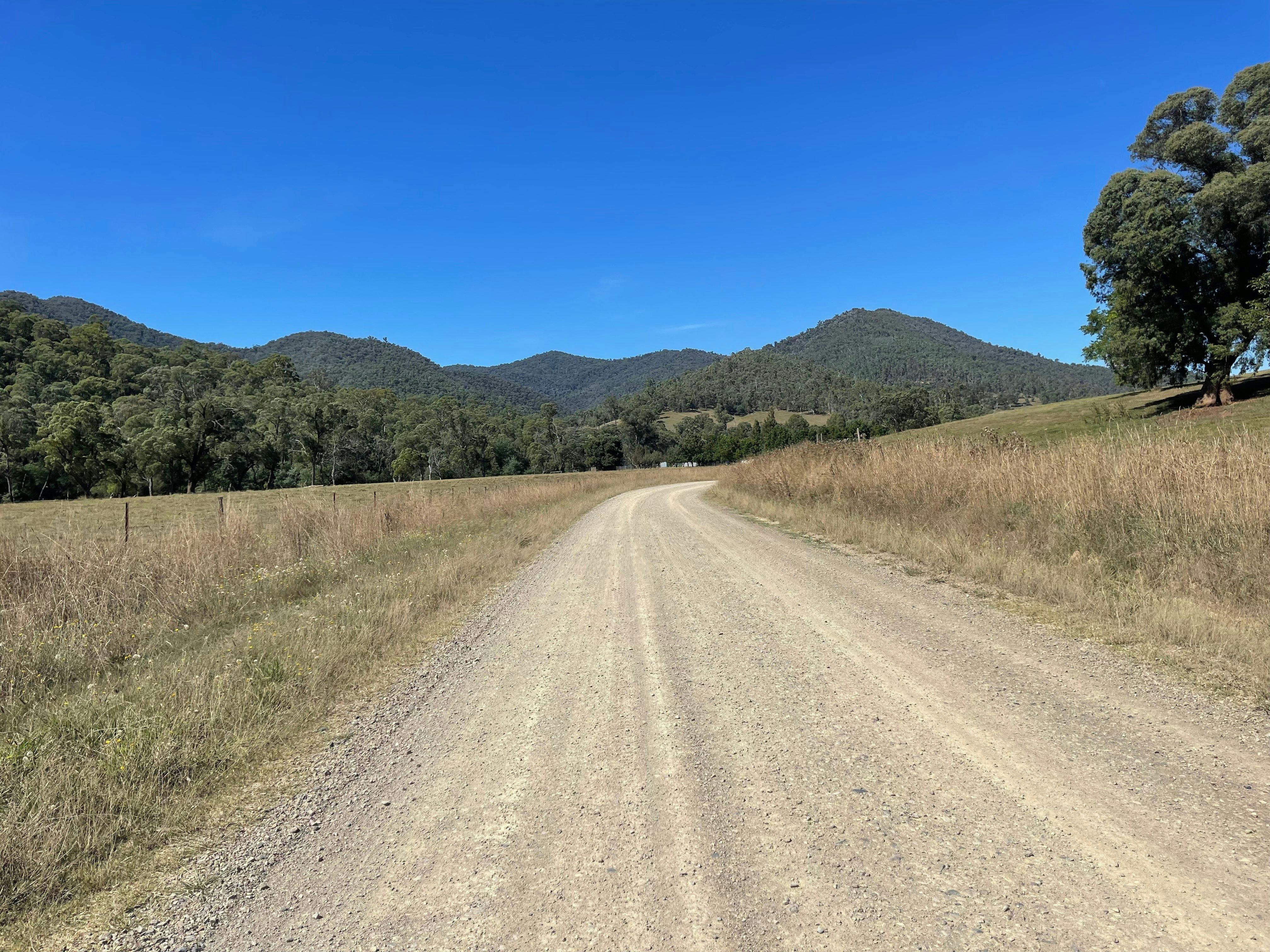 Gravel road with grey dirt, grass, hills, trees, blue sky