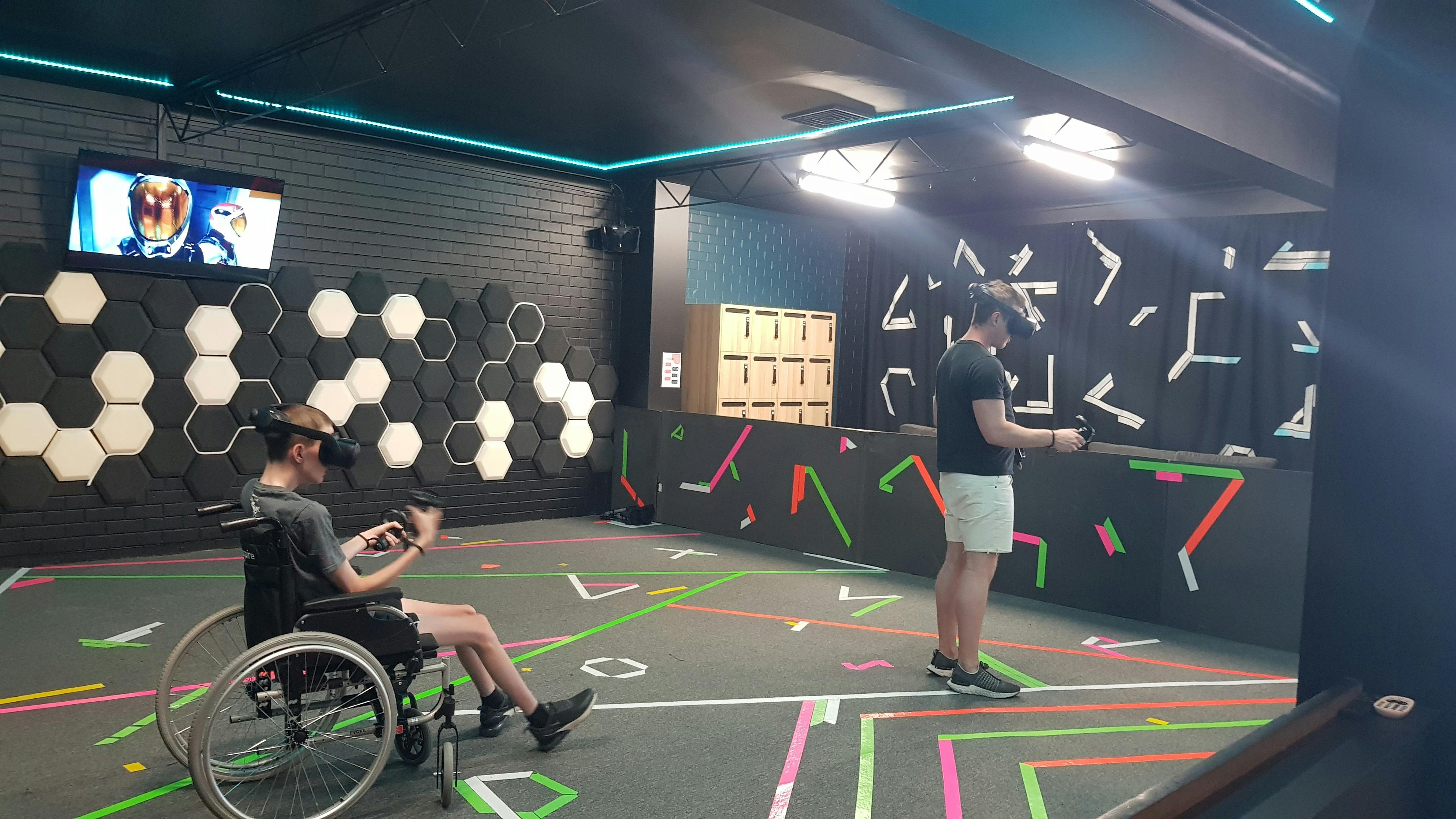 Two people VR headsets playing an action game, one of whom uses a wheelchair to aid mobility.