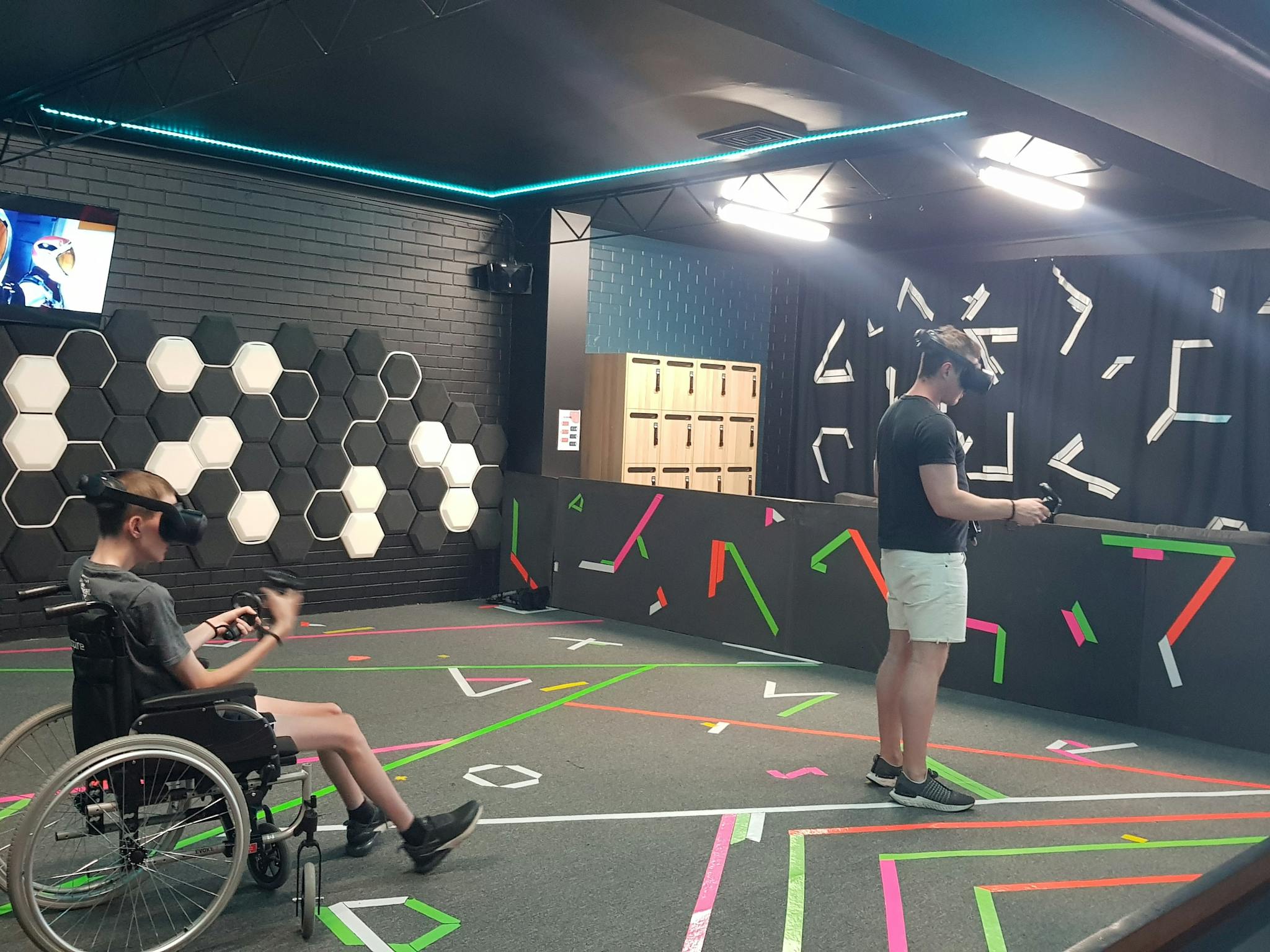 Two people VR headsets playing an action game, one of whom uses a wheelchair to aid mobility.