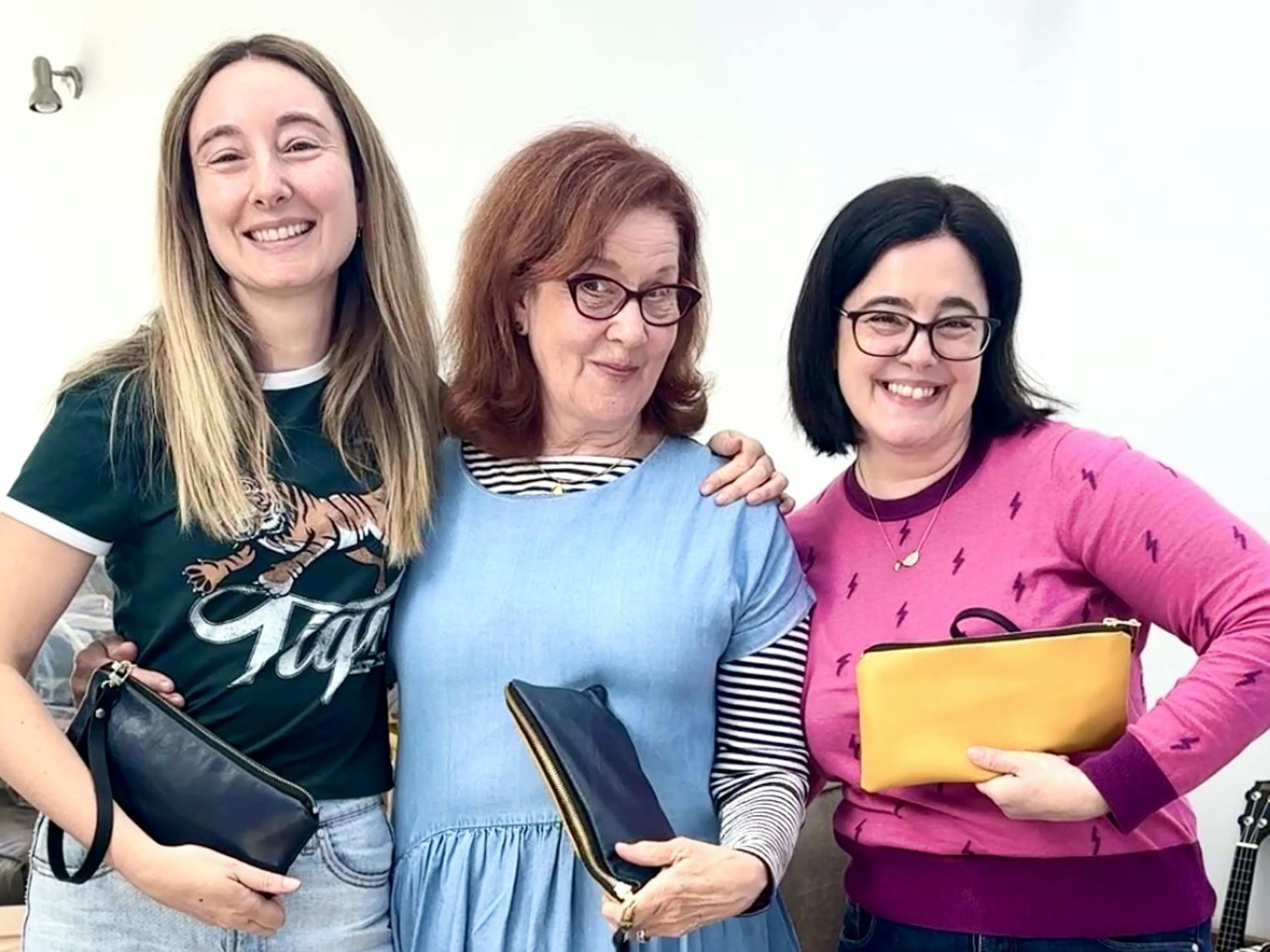 Three women smiling and proudly holding their handmade leather clutches from a workshop. The woman o