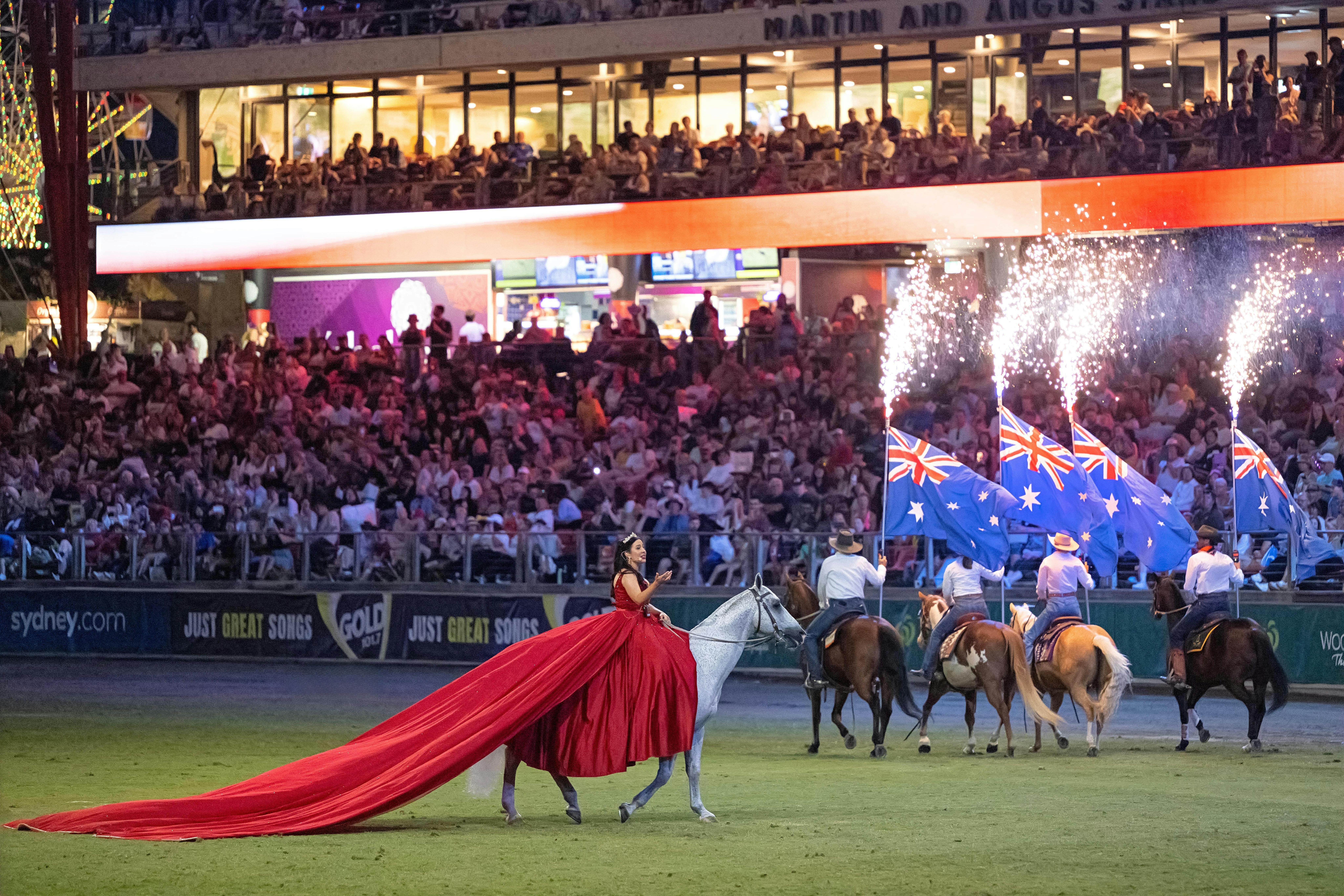 Woman in a red dress on horseback performing, with riders carrying Australian flags.