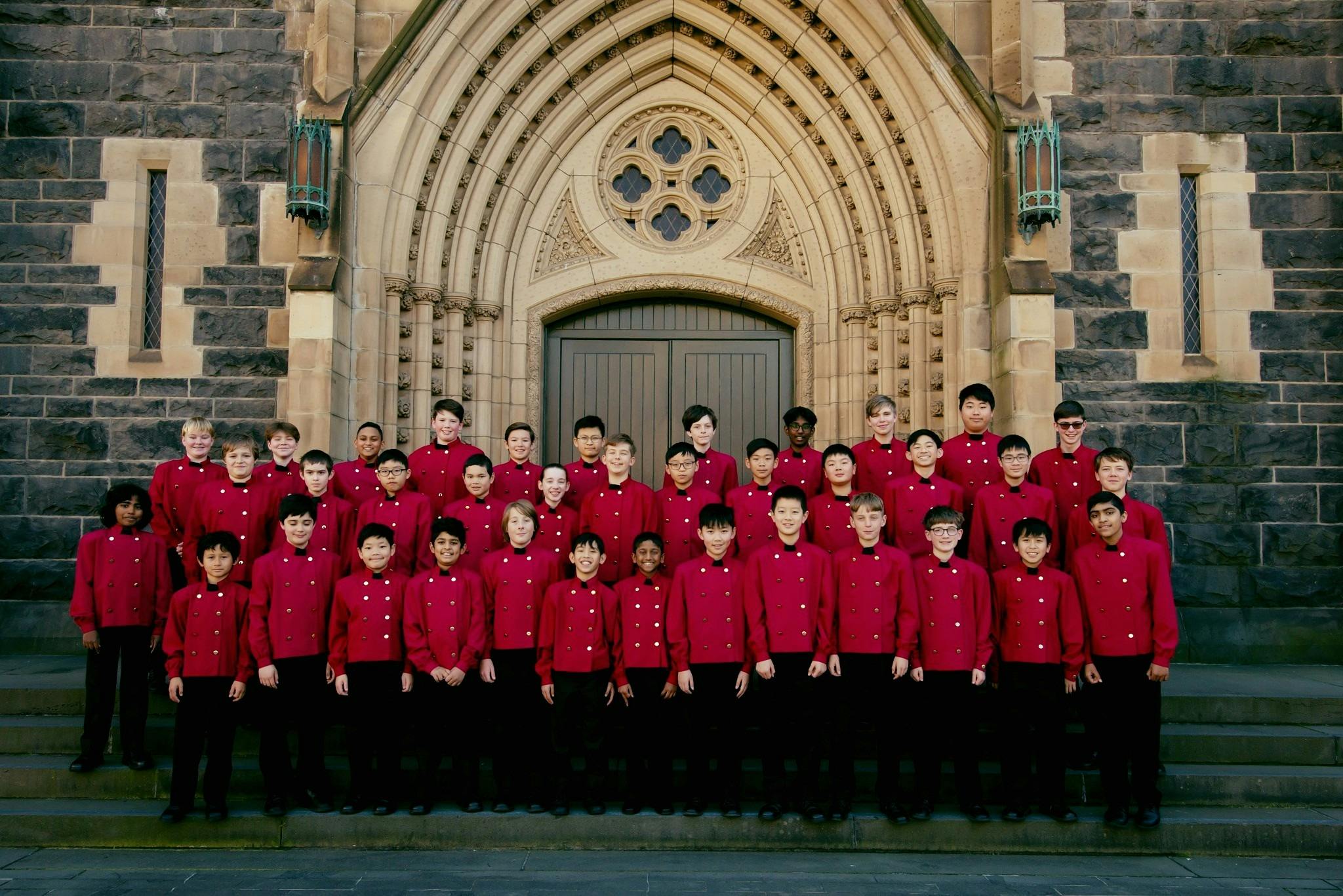 Boys choir stand together wearing red coats in front of bluestone cathedral
