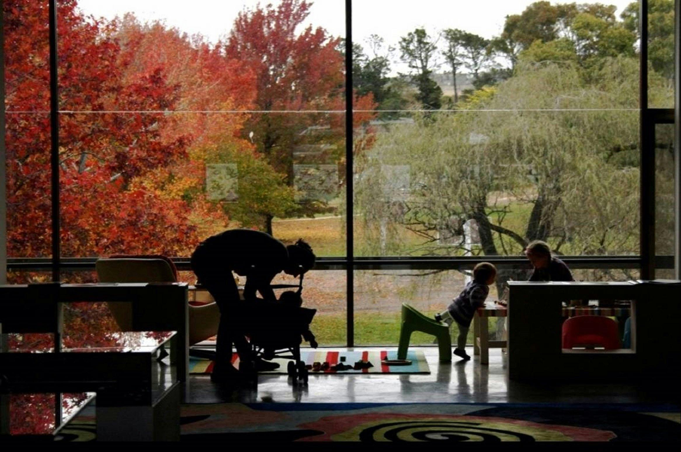 Two adults with two children playing with toys in front of a big window overlooking a leafy parkland