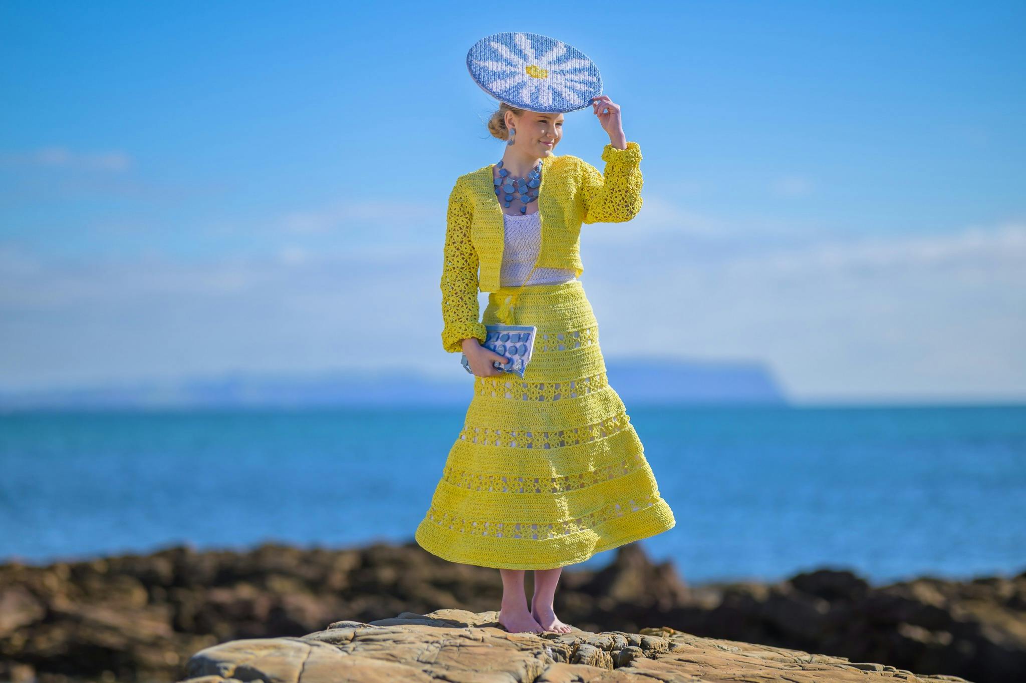 Model wearing brightly coloured yellow outfit with a daisy-patterned hat in a beachside setting