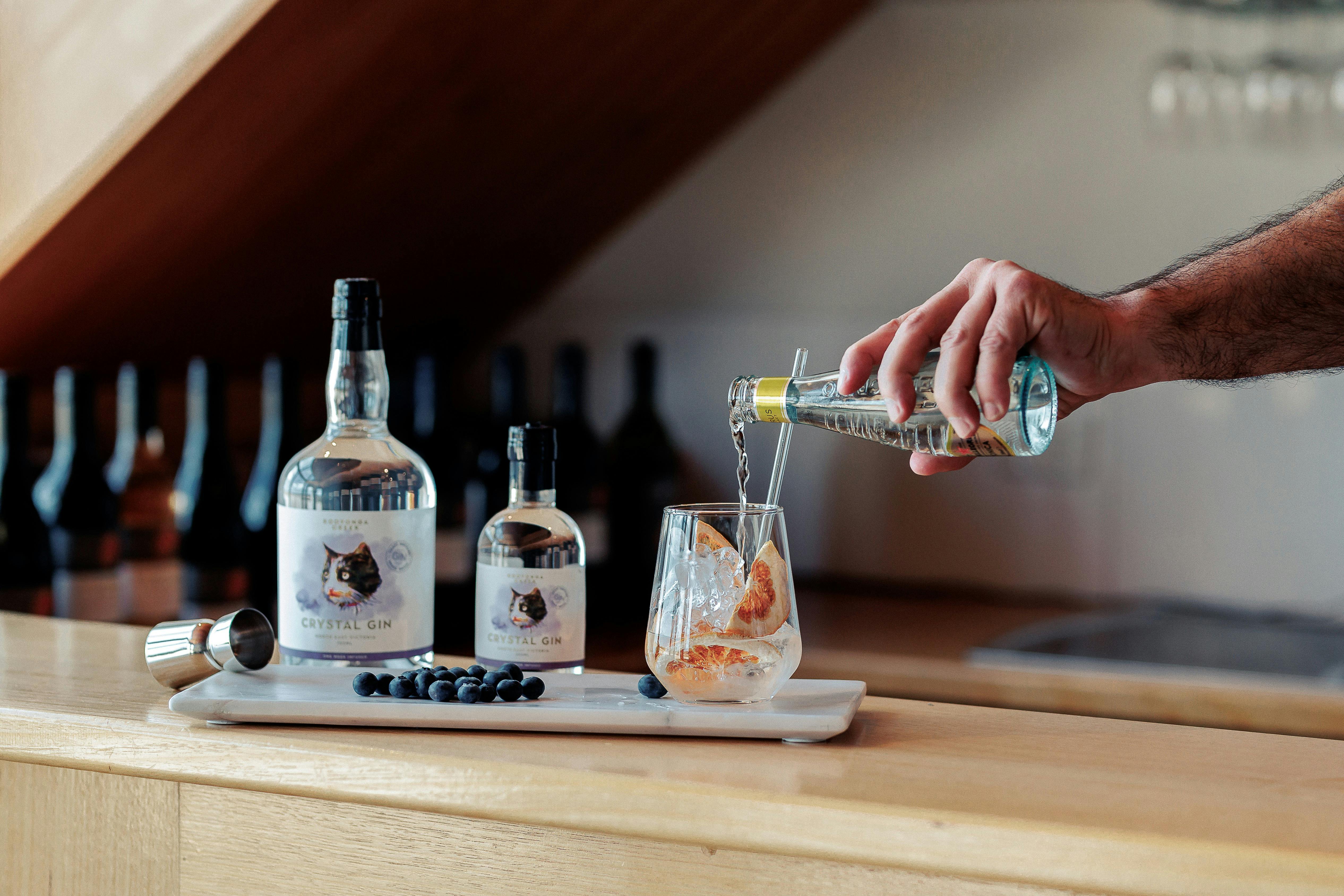 Person pouring tonic into a glass with ice and  Crystal Gin beside a small and large Gin  bottle