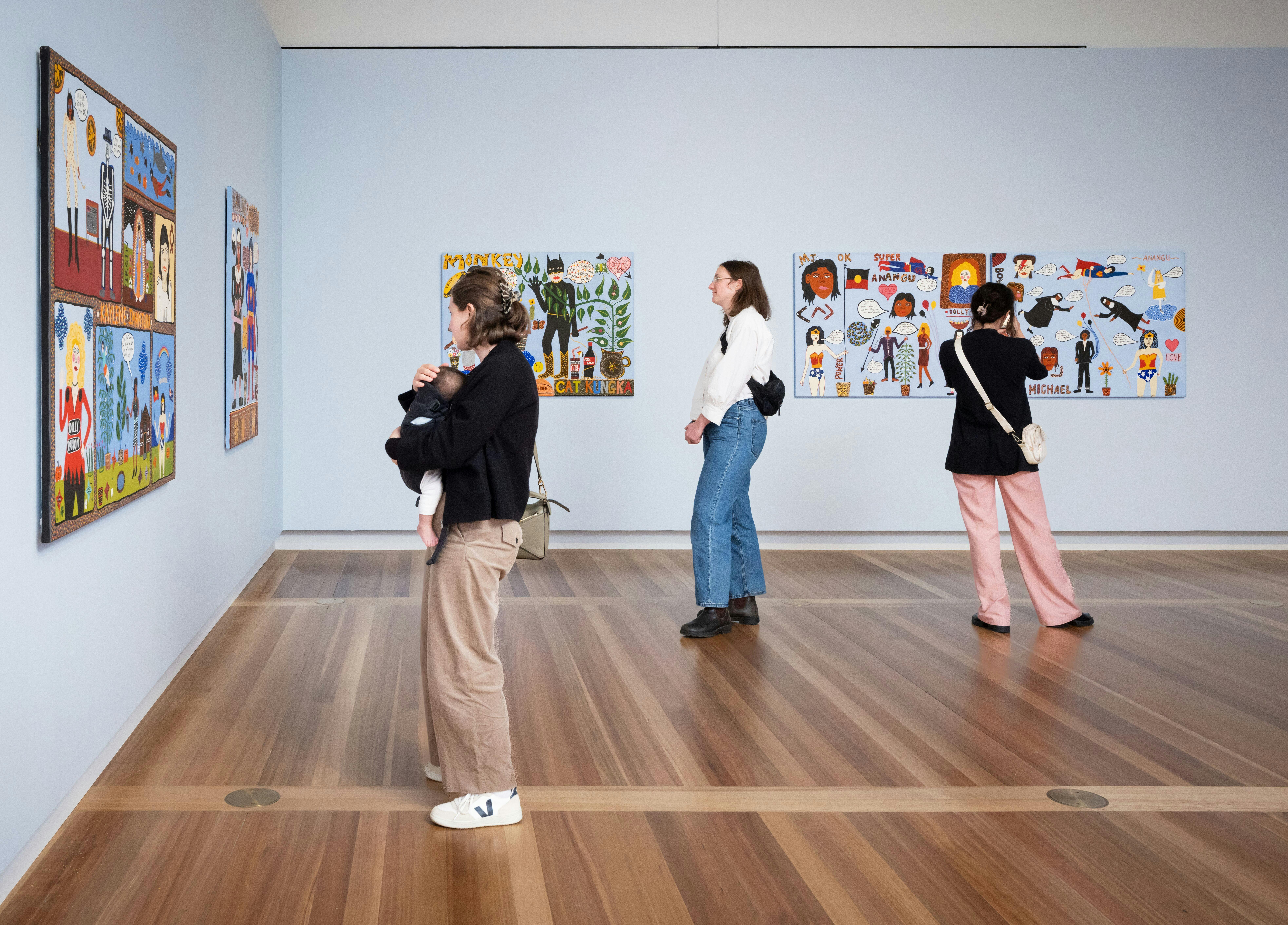 Three people look at colourful portraits hanging on pale blue walls. One of them holds a baby.