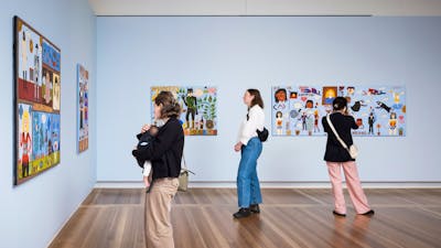 Three people look at colourful portraits hanging on pale blue walls. One of them holds a baby.