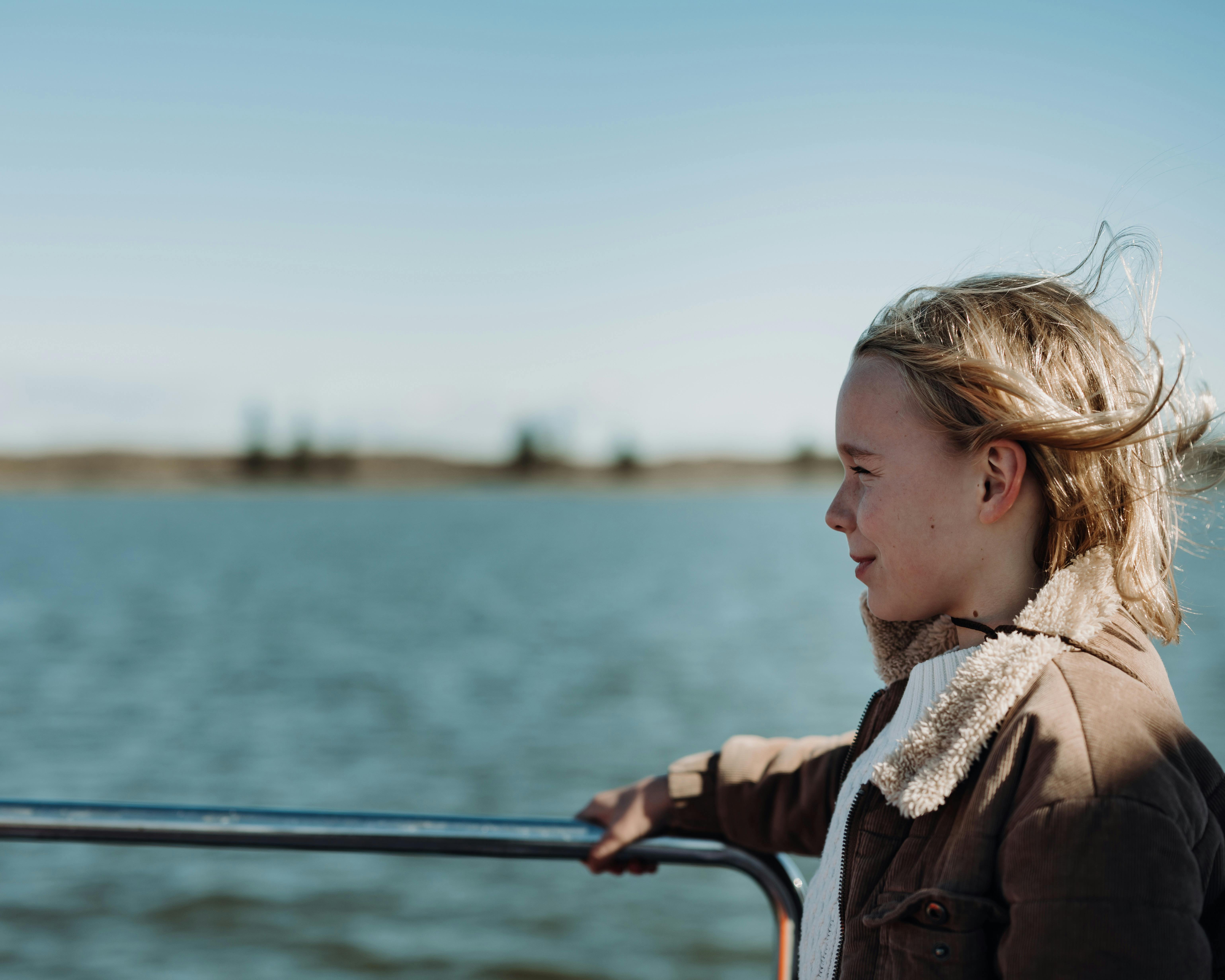 A child in a brown jacket enjoys a boat ride, gazing at the water with windblown hair.