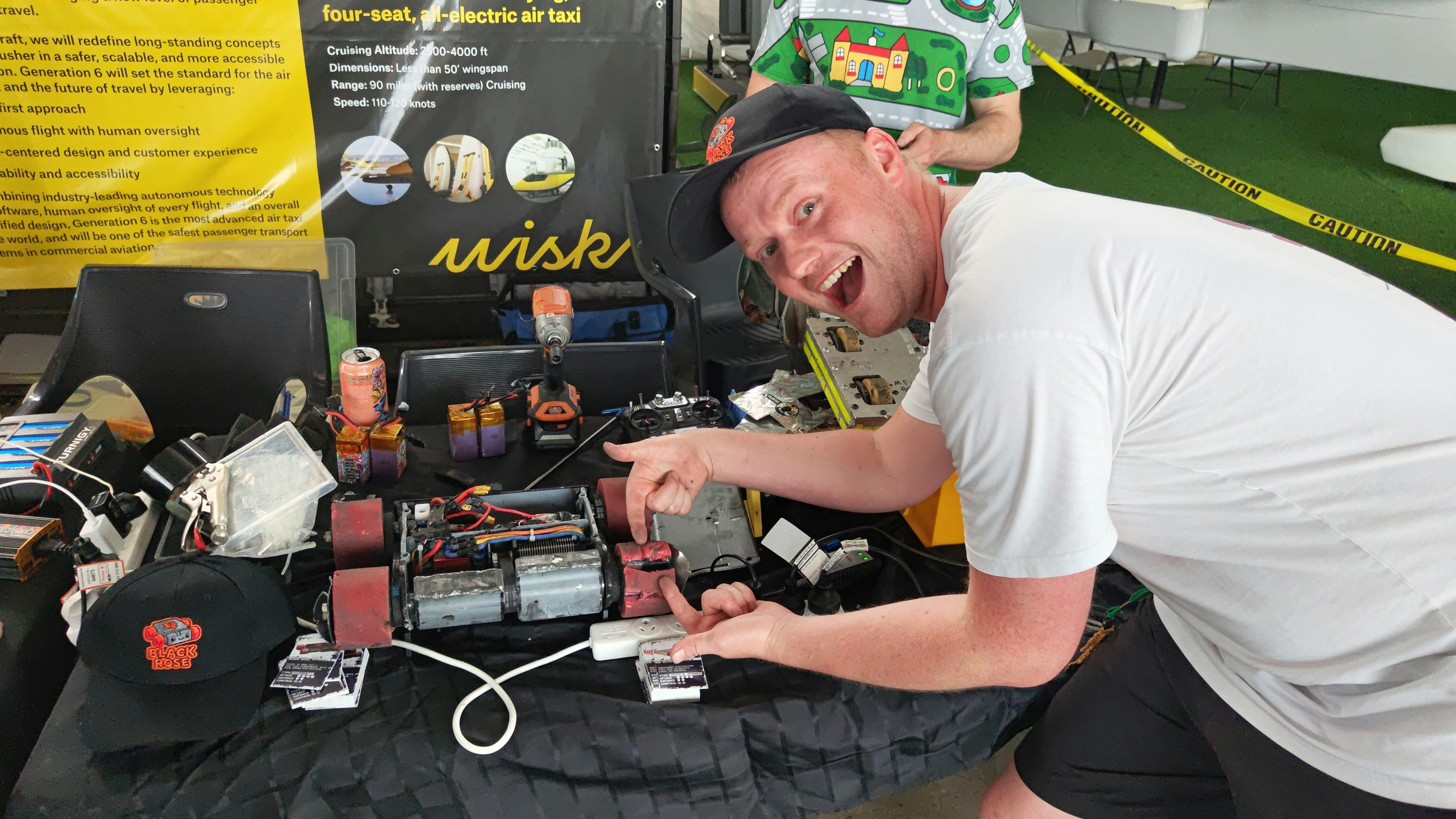 Robot builder beside an open combat robot, tools and radio controllers in the Robowars pits.