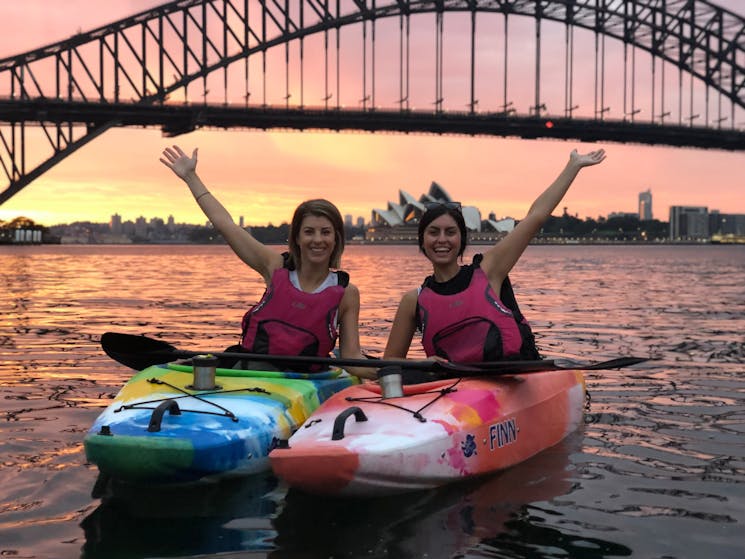 Two girls in single kayaks on Sydney Harbour with a pink sunrise in front of the Opera House