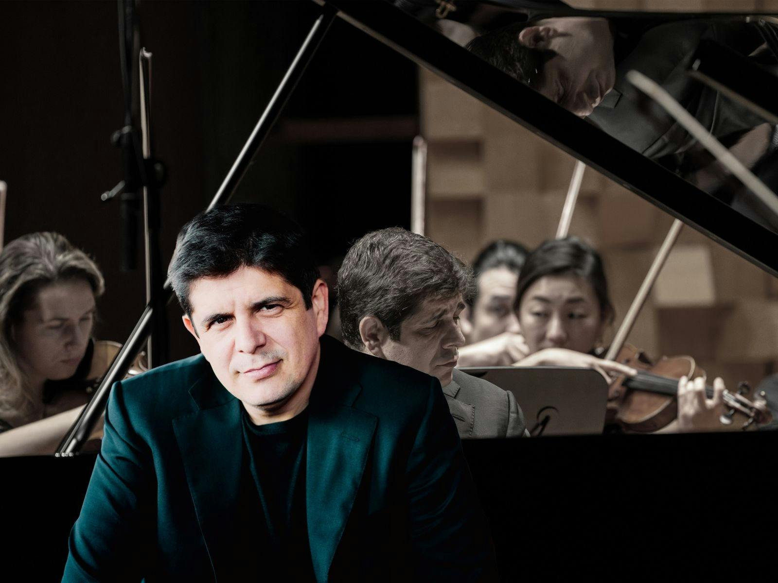 A man sitting in front of a concert piano, with violin players in the background.