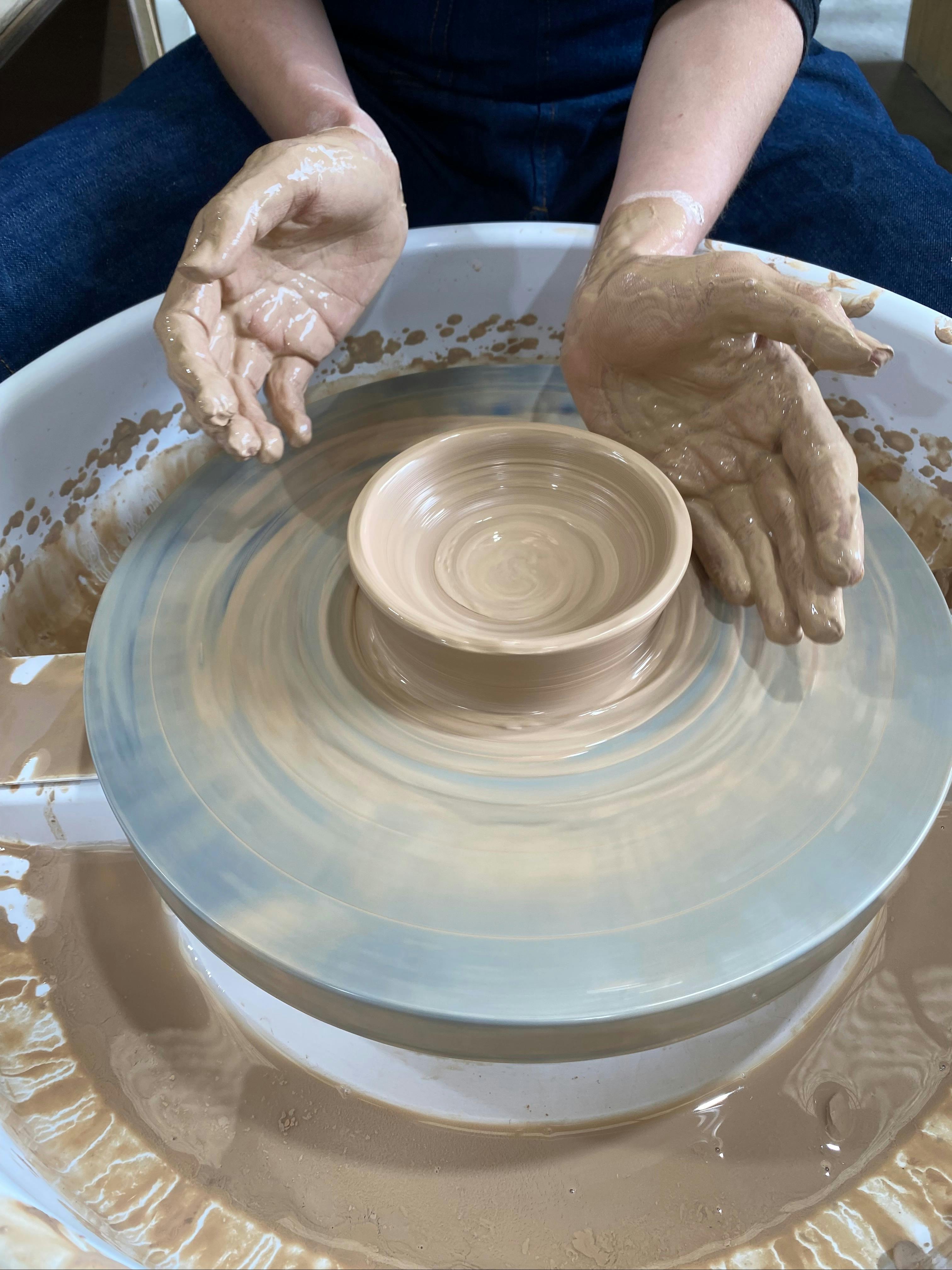 hands covered in clay around a clay vessel on a pottery wheel