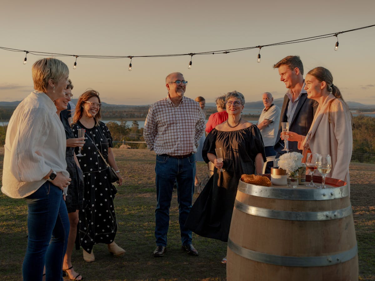 a group of people standing in a country field with wine barrels and fairy lights enjoying  food